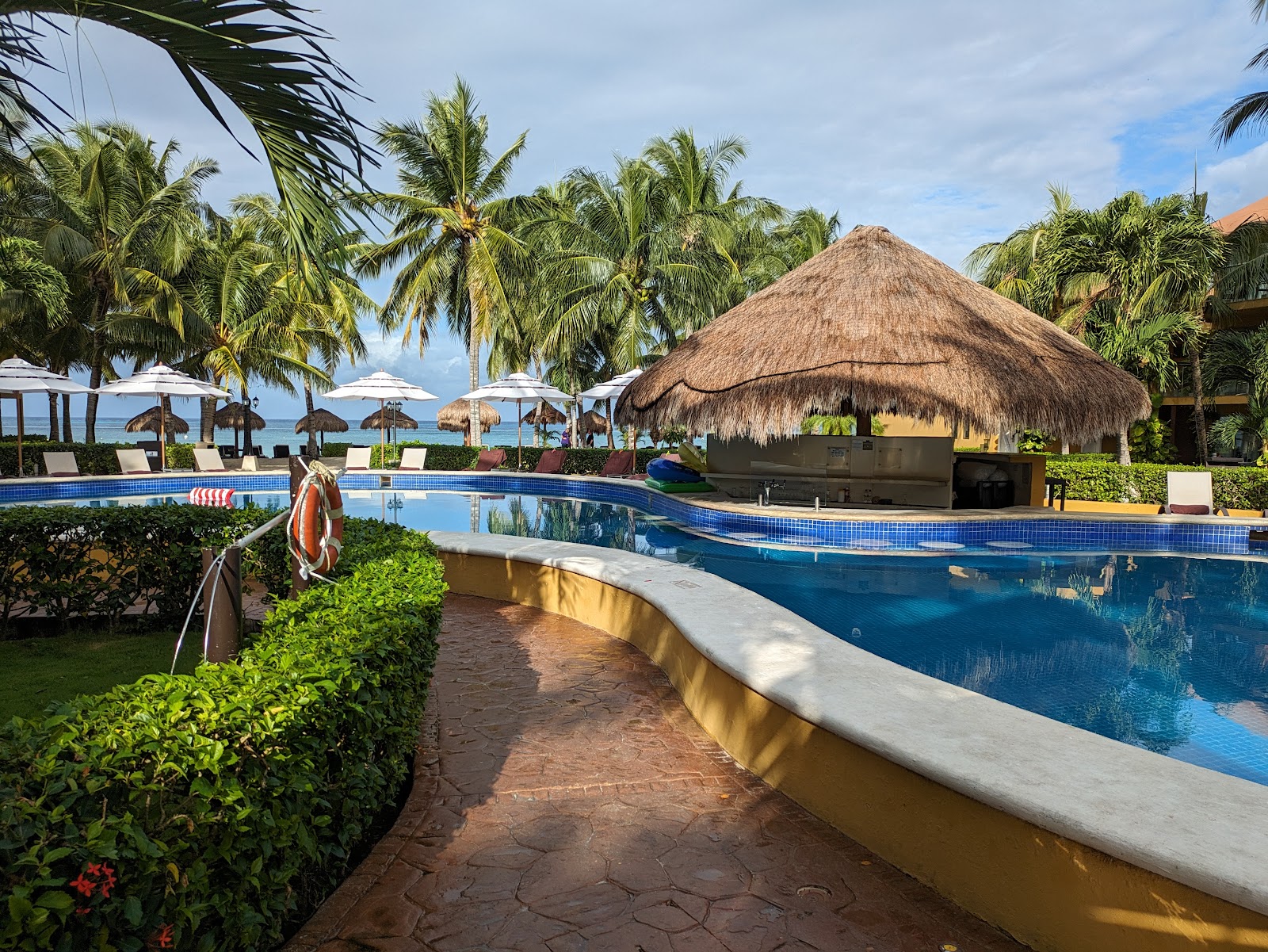 Curved pool with blue tile borders overlooking the ocean, flanked by palm trees and a thatched-roof structure at Secrets Aura Cozumel.
