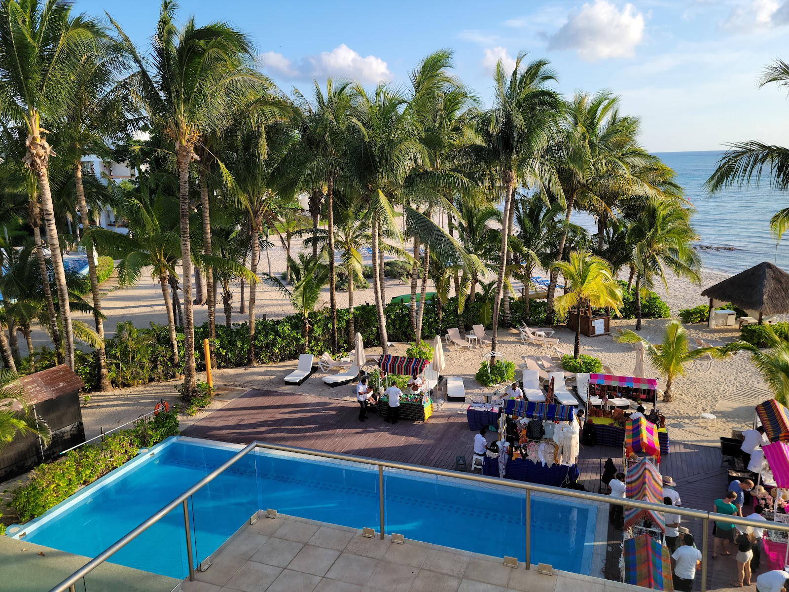 Blue pool in foreground overlooks palm-lined beach with lounge chairs, thatched-roof bar, and ocean at Secrets Aura Cozumel.