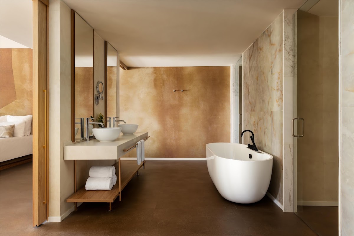 Modern bathroom with white soaking tub, vessel sink vanity, warm travertine walls, and walk-in shower at Secrets Baby Beach Aruba.