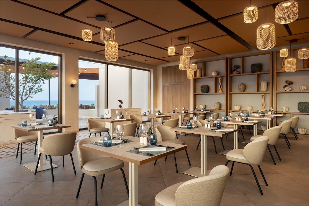 Dining room with beige chairs at wood tables, woven pendant lights overhead, ocean view through floor-to-ceiling windows, and wooden shelving displaying decorative objects.