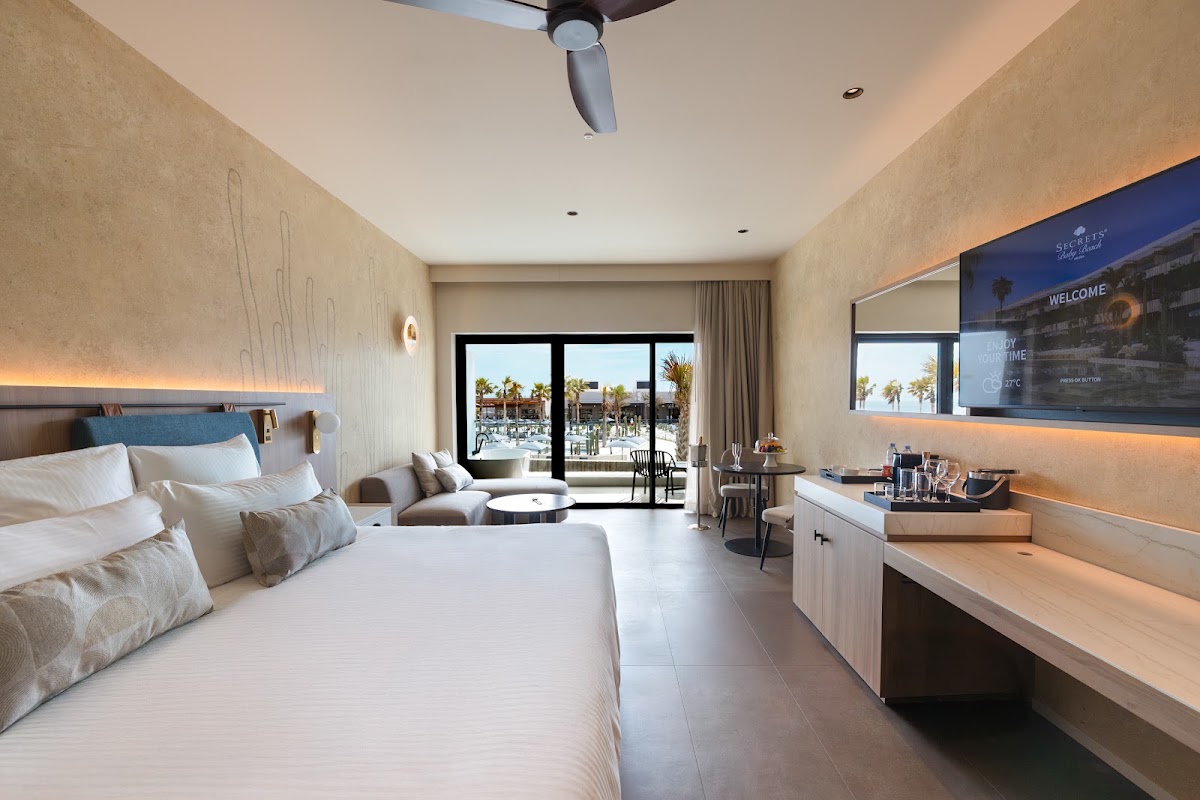 Modern resort bedroom with king bed, vanity desk, and balcony overlooking pool and palm trees at Secrets Baby Beach Aruba.