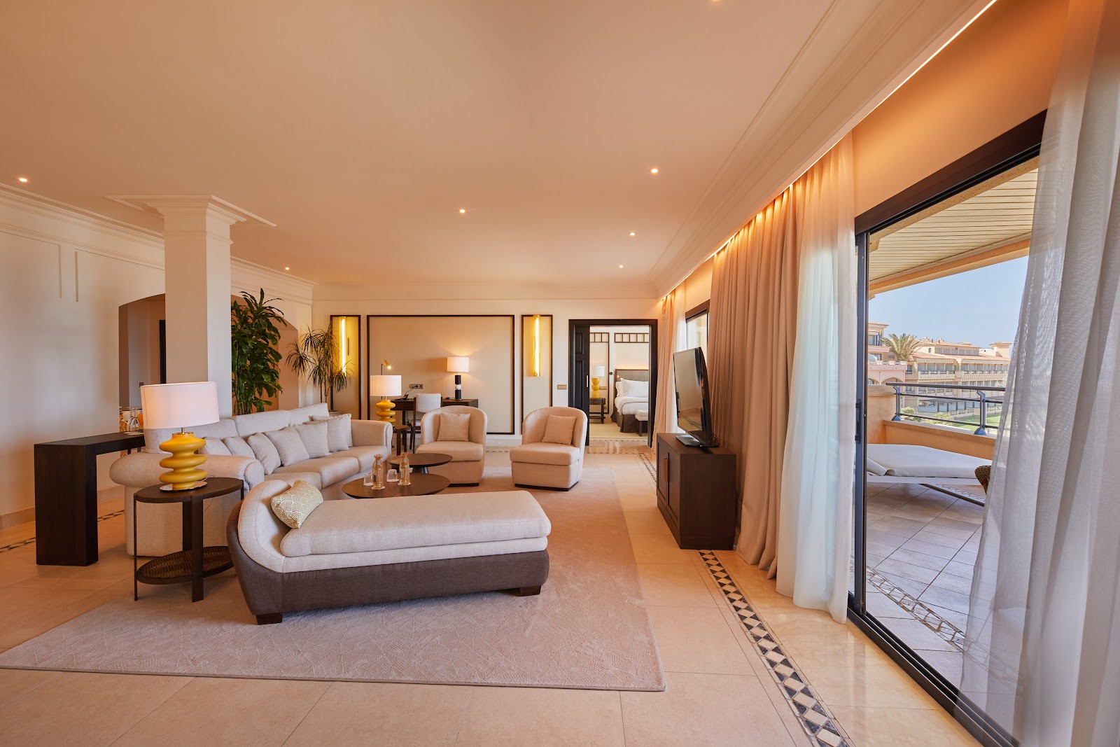 Spacious suite living room with cream furnishings, lit ceiling, and glass doors opening to a balcony overlooking a coastal resort.