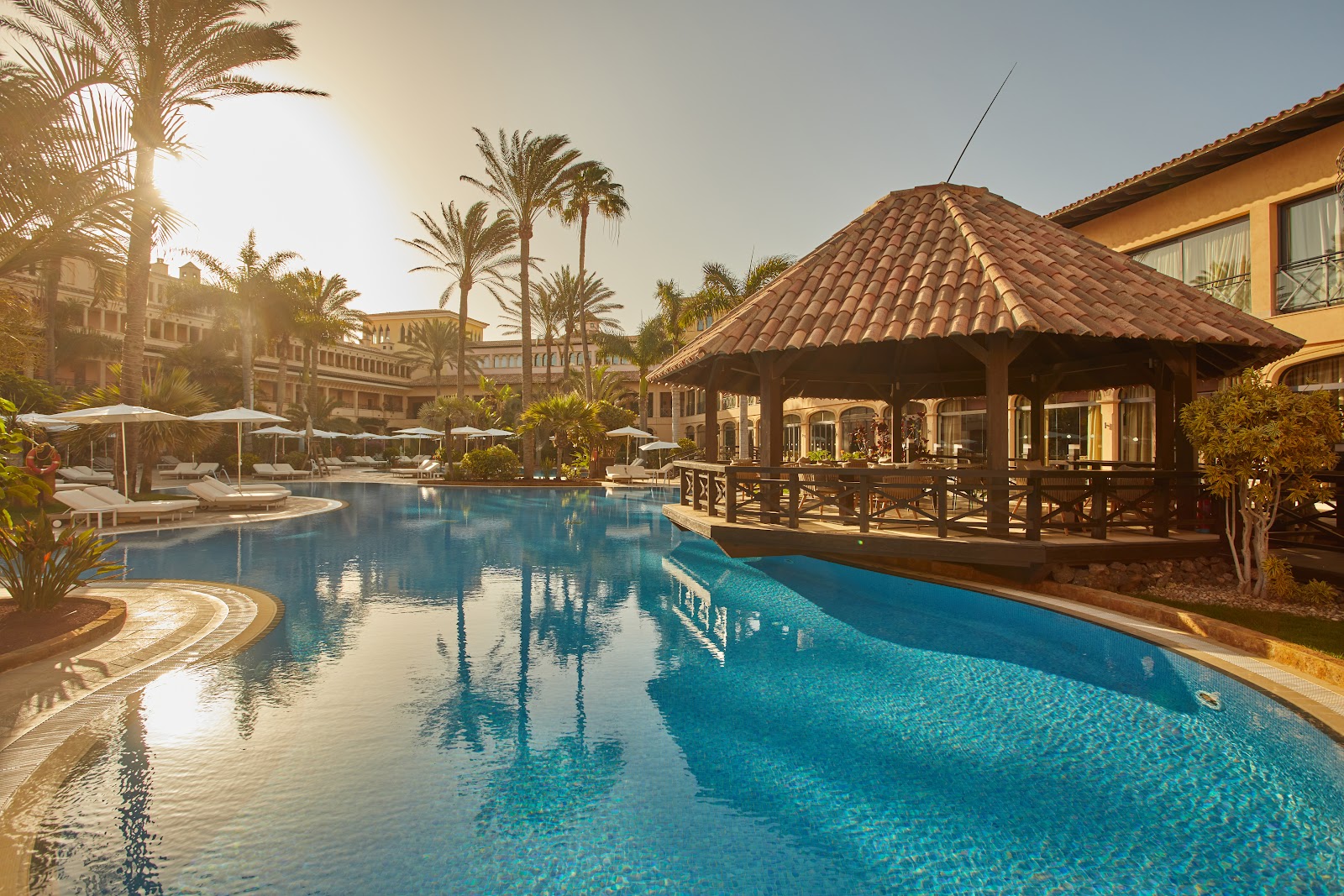 Resort pool with blue water at sunset, flanked by a tiled-roof gazebo, palm trees, white umbrellas, and golden-colored buildings.