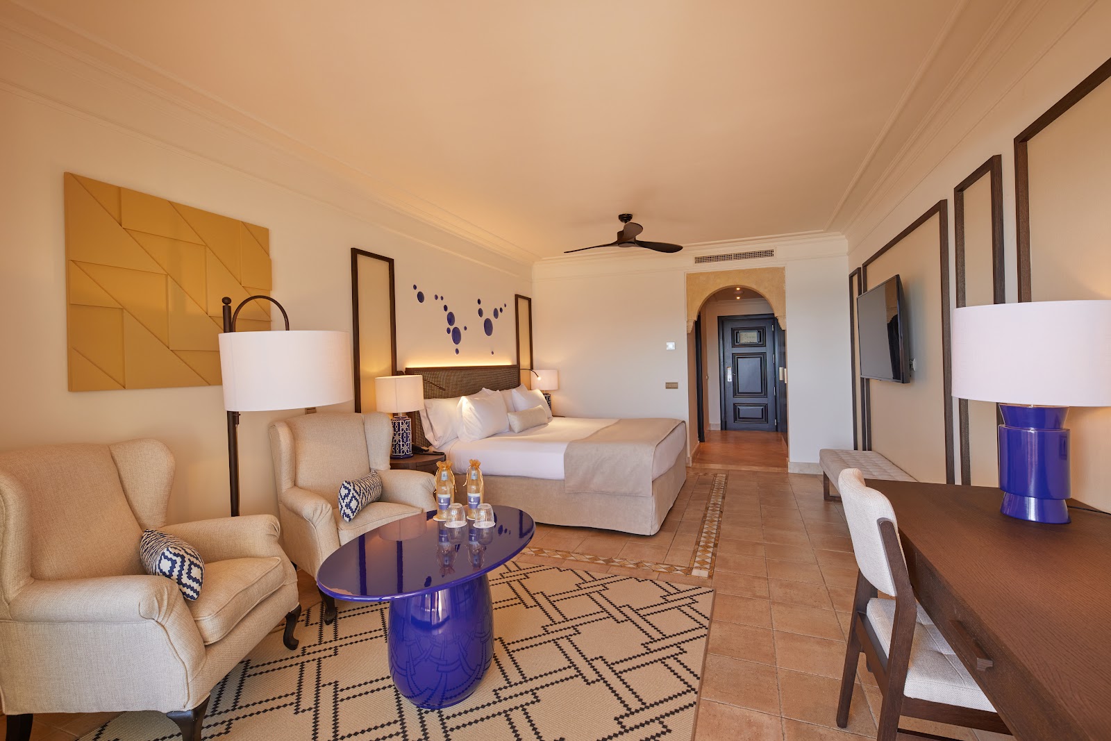 Spacious resort suite with cream upholstery seating, blue accent table, king bed with headboard lighting, and terracotta tile floors.