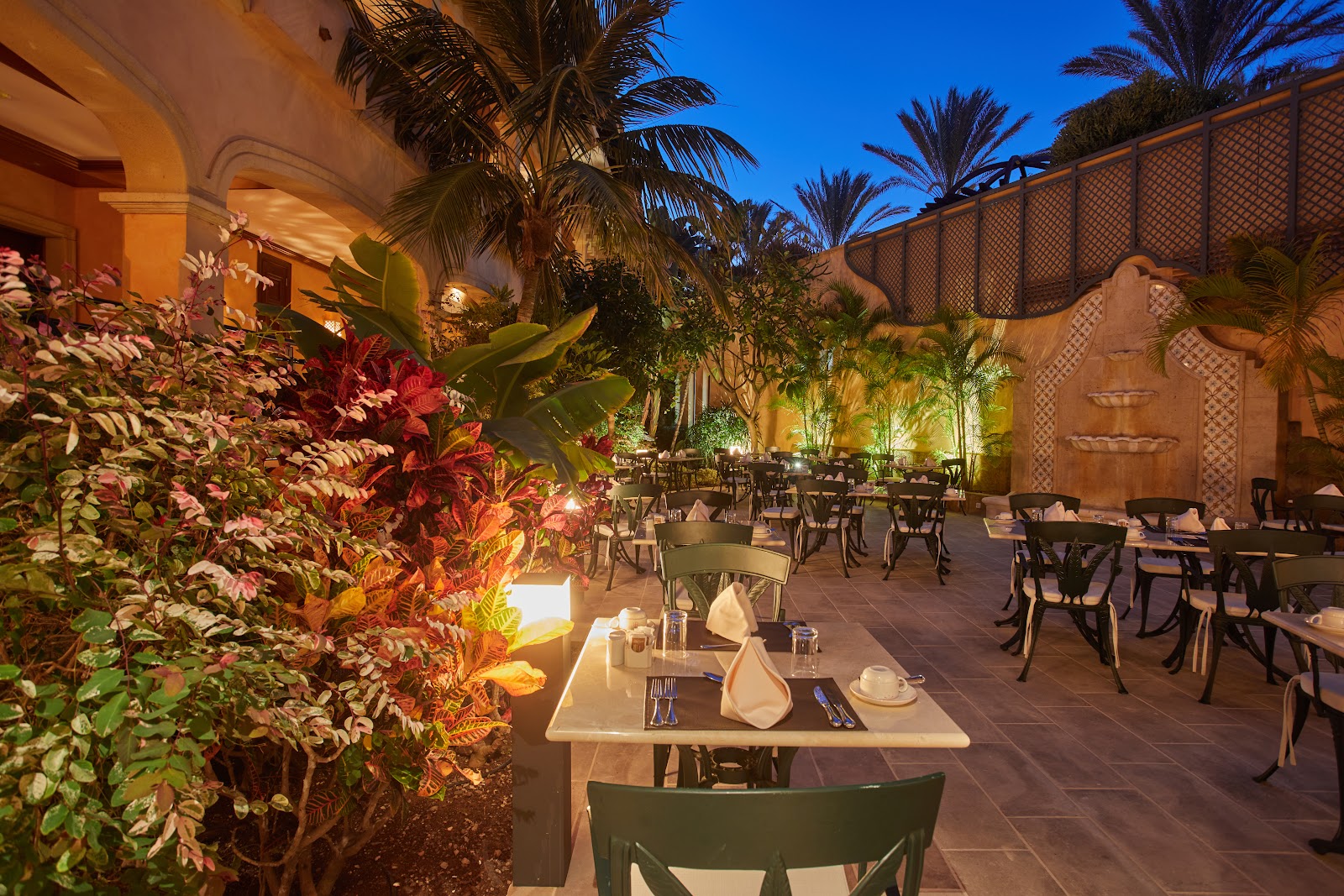 Outdoor dining terrace at dusk with set tables, palm trees, flowering plants, and illuminated ochre-colored building at Secrets Bahia Real Resort & Spa.