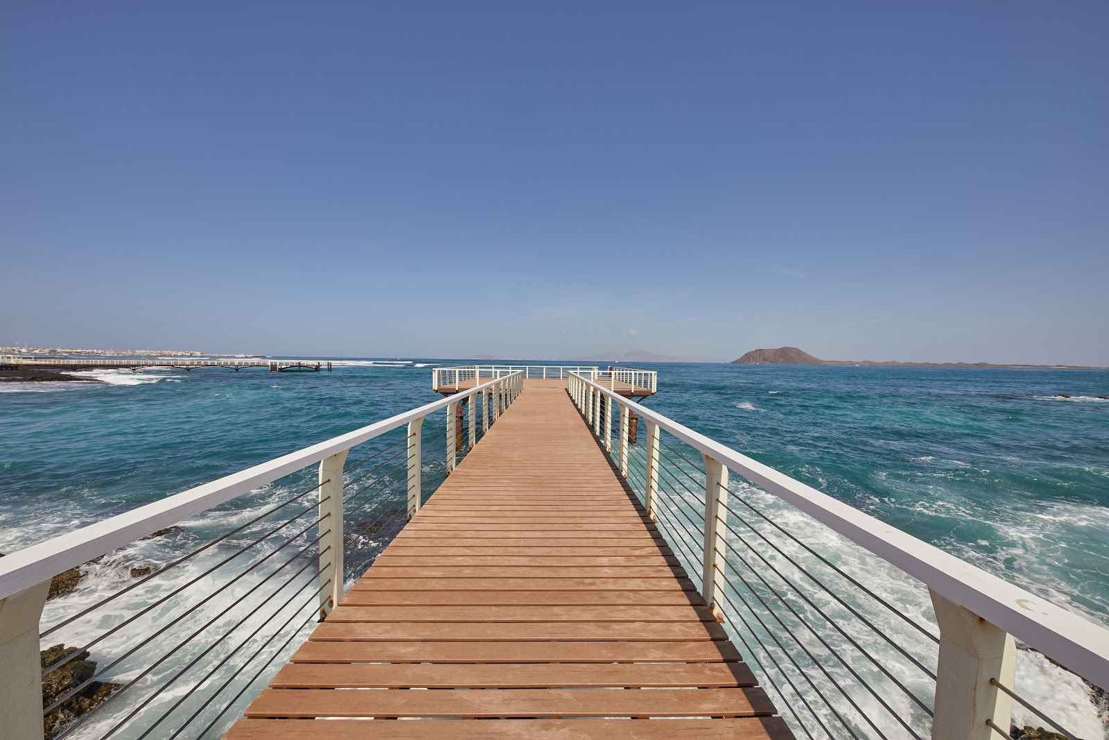 Wooden pier with white railings extending into turquoise ocean waters at Secrets Bahia Real Resort & Spa, with a volcanic island visible on the horizon.