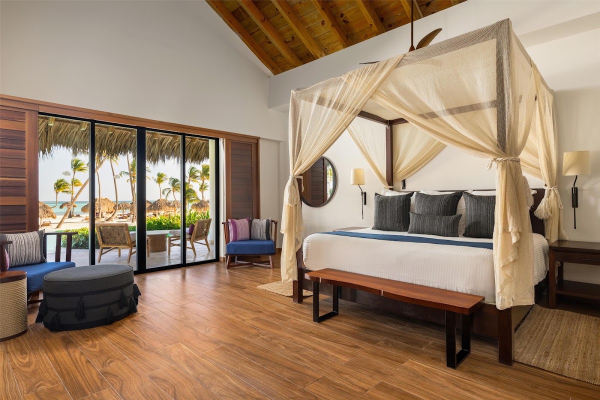 Spacious bedroom suite with four-poster canopy bed, wooden beams, and ocean view through sliding glass doors with palm trees visible.