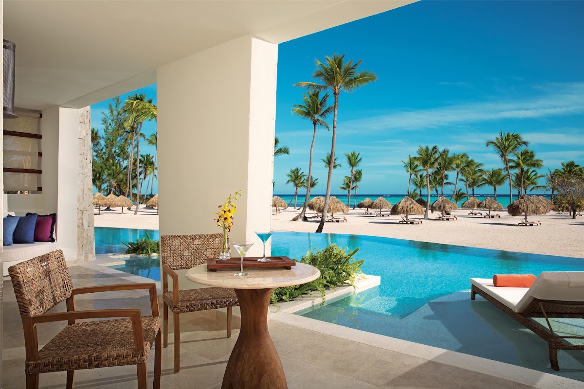 Resort lounge with woven chairs overlooking an infinity pool, beach with palm trees, and turquoise ocean beyond.