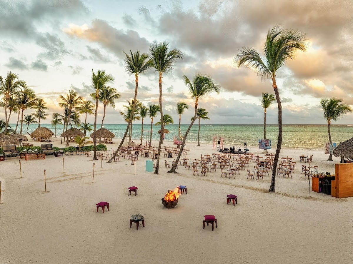 Beach setup at Secrets Cap Cana with palm trees, burgundy seating, a lit fire pit, thatched palapas, and turquoise ocean under cloudy sky.