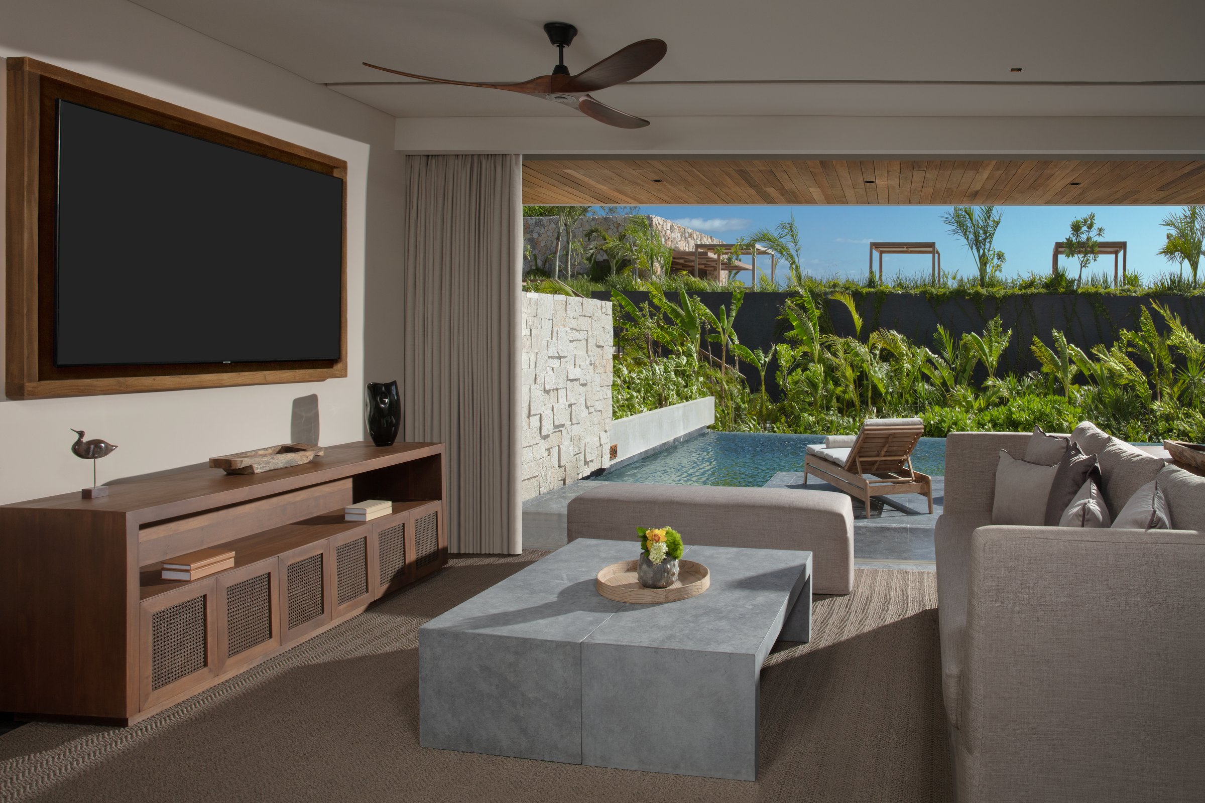 Master swim-out suite living room with a gray sectional, wood console and glass doors to a terrace with a plunge pool and greenery.