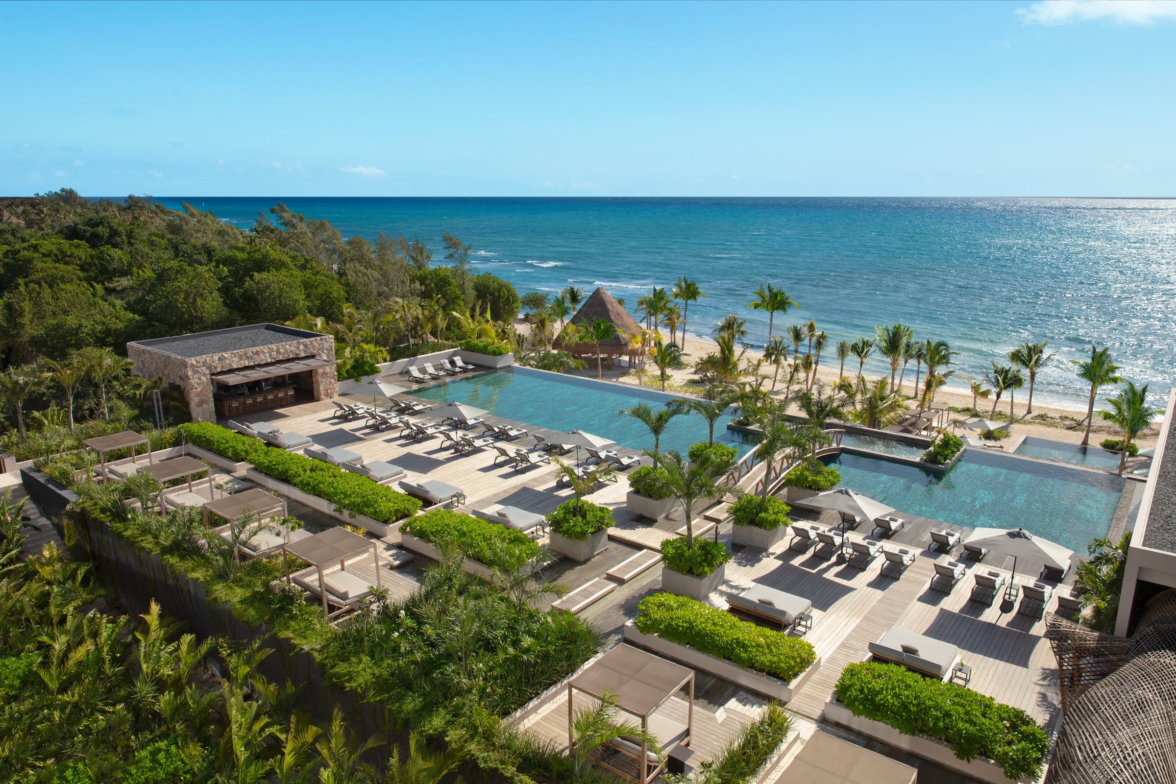 High-angle view of the main courtyard pools with rows of loungers and palm landscaping, with the ocean beyond.