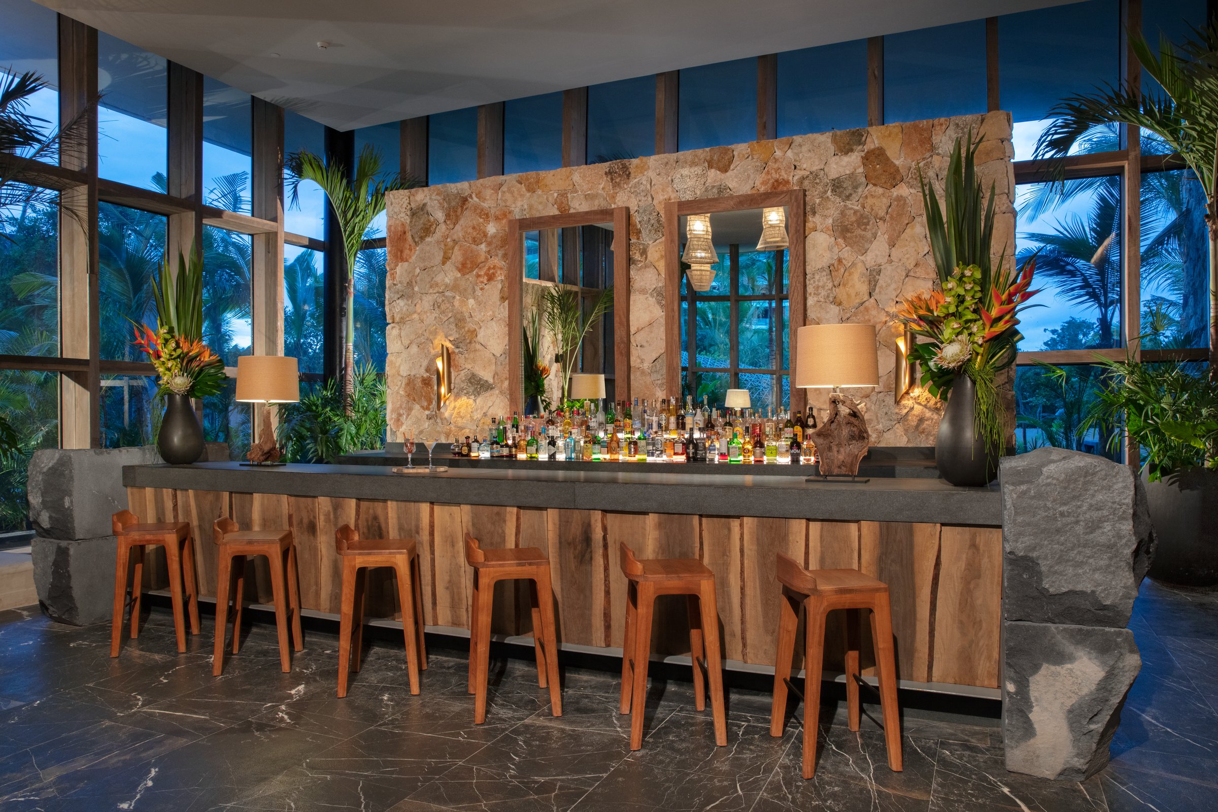 Lobby bar with a dark counter, warm wood and stone base, wood stools and a textured stone back wall flanked by large floral arrangements.