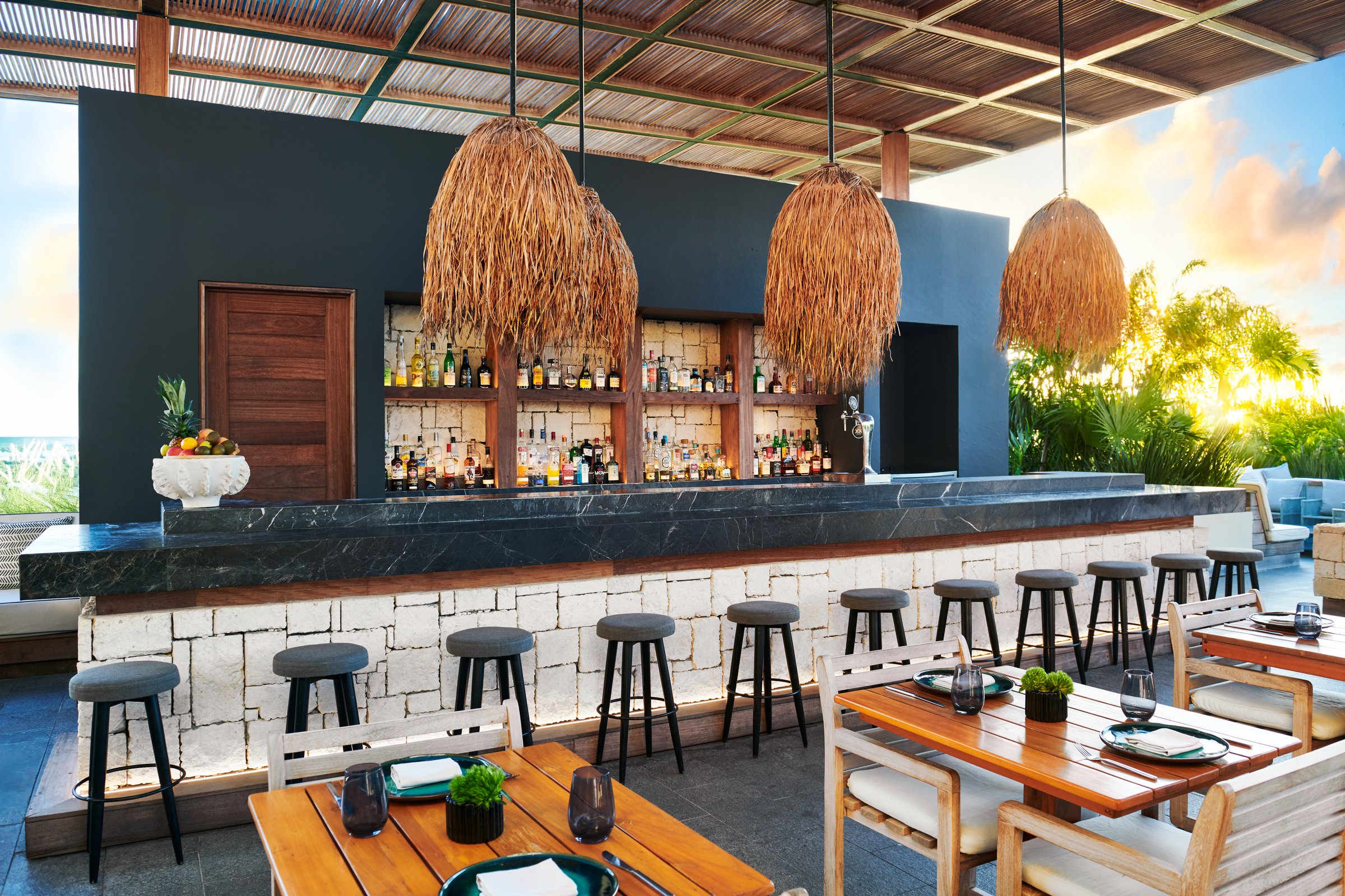 Cielo outdoor bar with a black marble top, white tiled front and dark wood stools under woven pendant lights and a slatted wood ceiling.