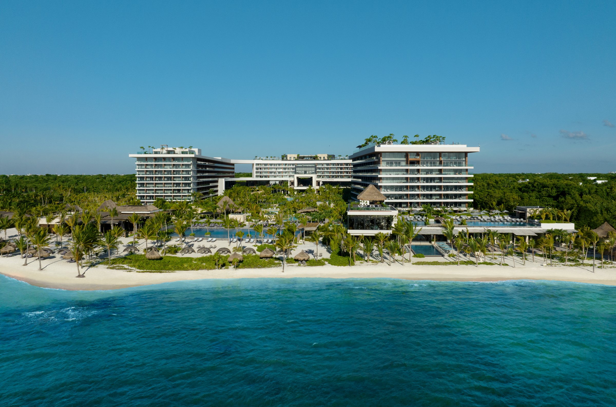 Aerial view of the multi-story white resort buildings along the white sand beach and turquoise sea, with pools among the palms.