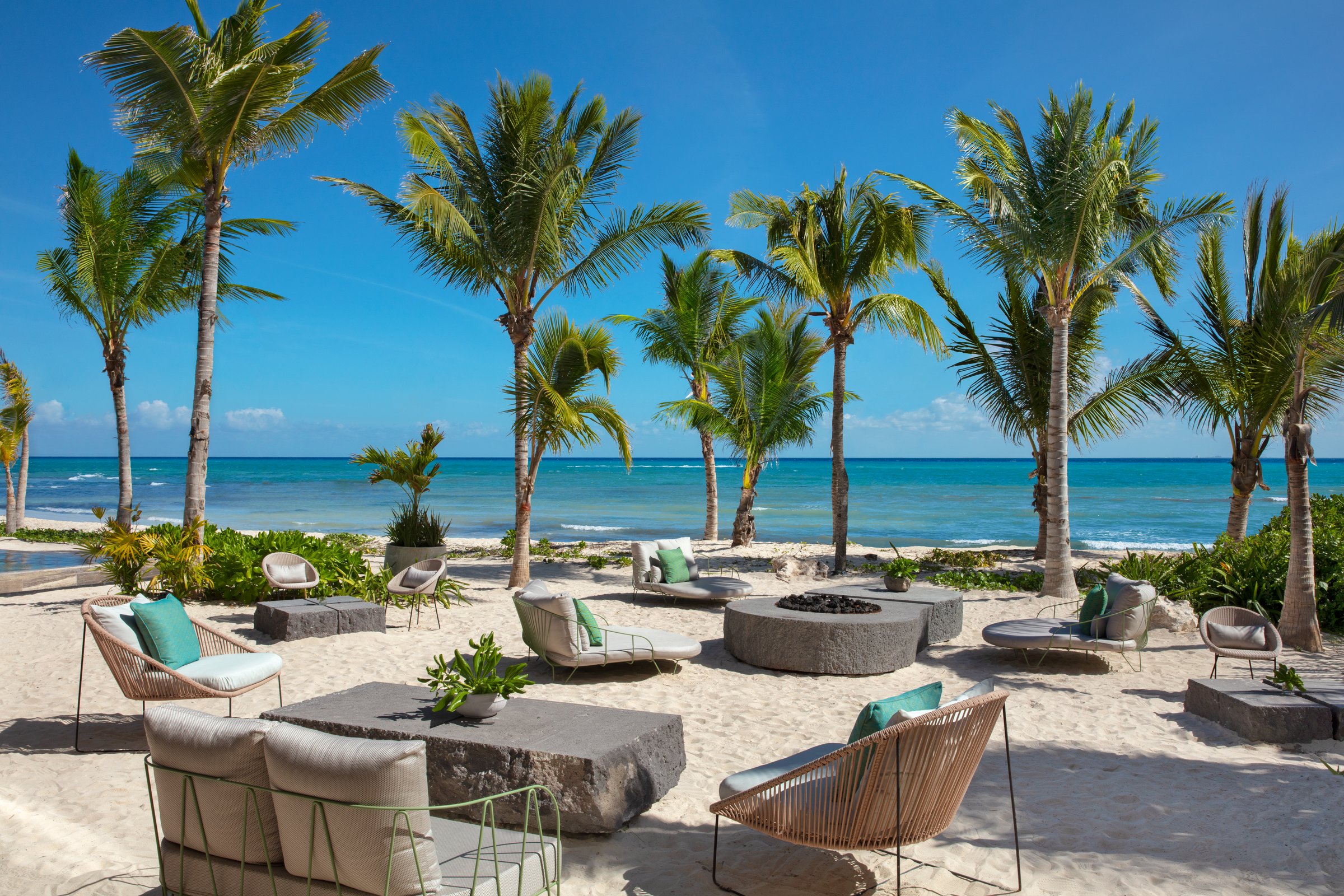 Beach club lounge on the sand with low modern sofas, stone coffee tables and a fire pit under palm trees and a blue sky.