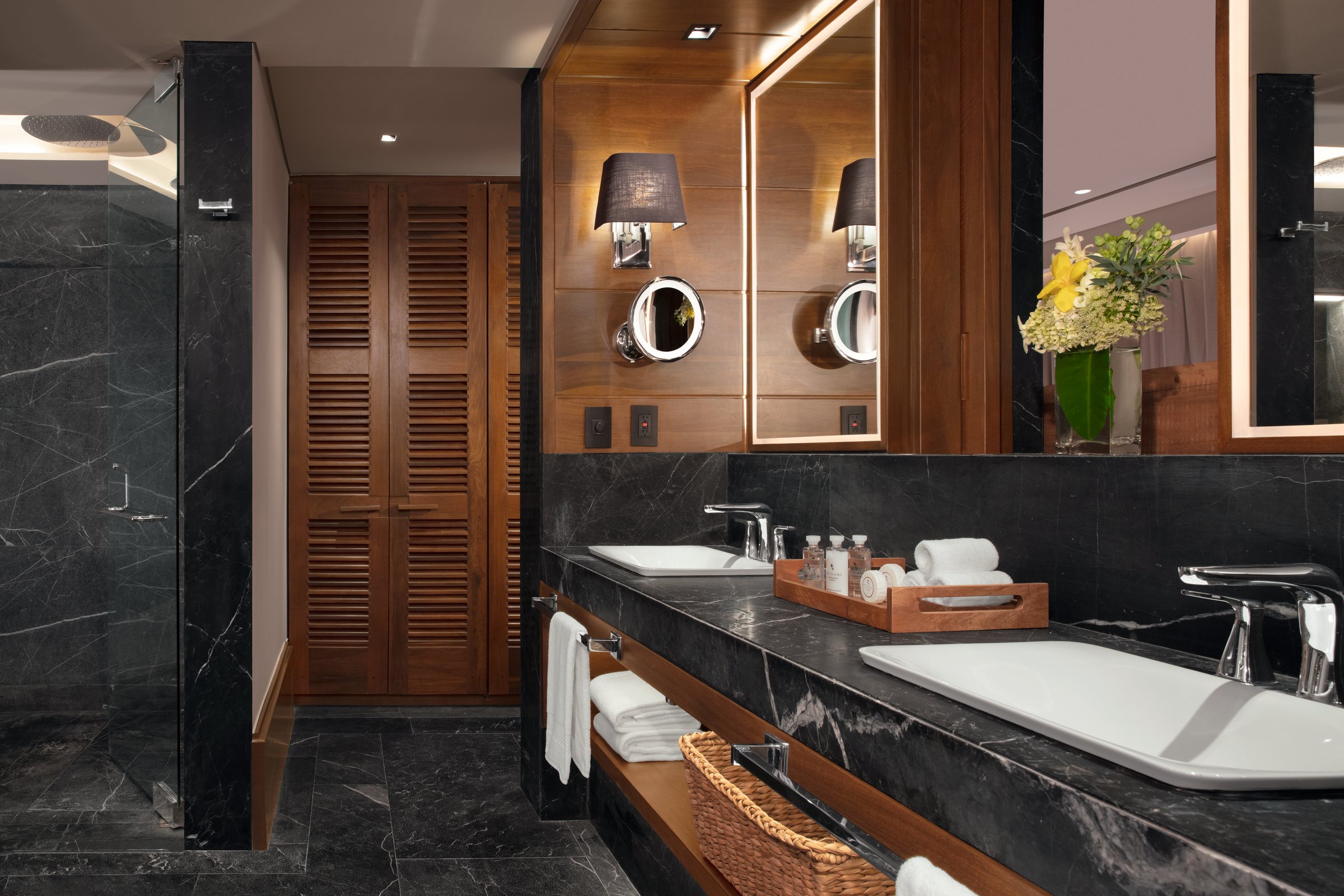 Suite bathroom with a long dark marble vanity, two white sinks, backlit mirrors and a walk-in shower.