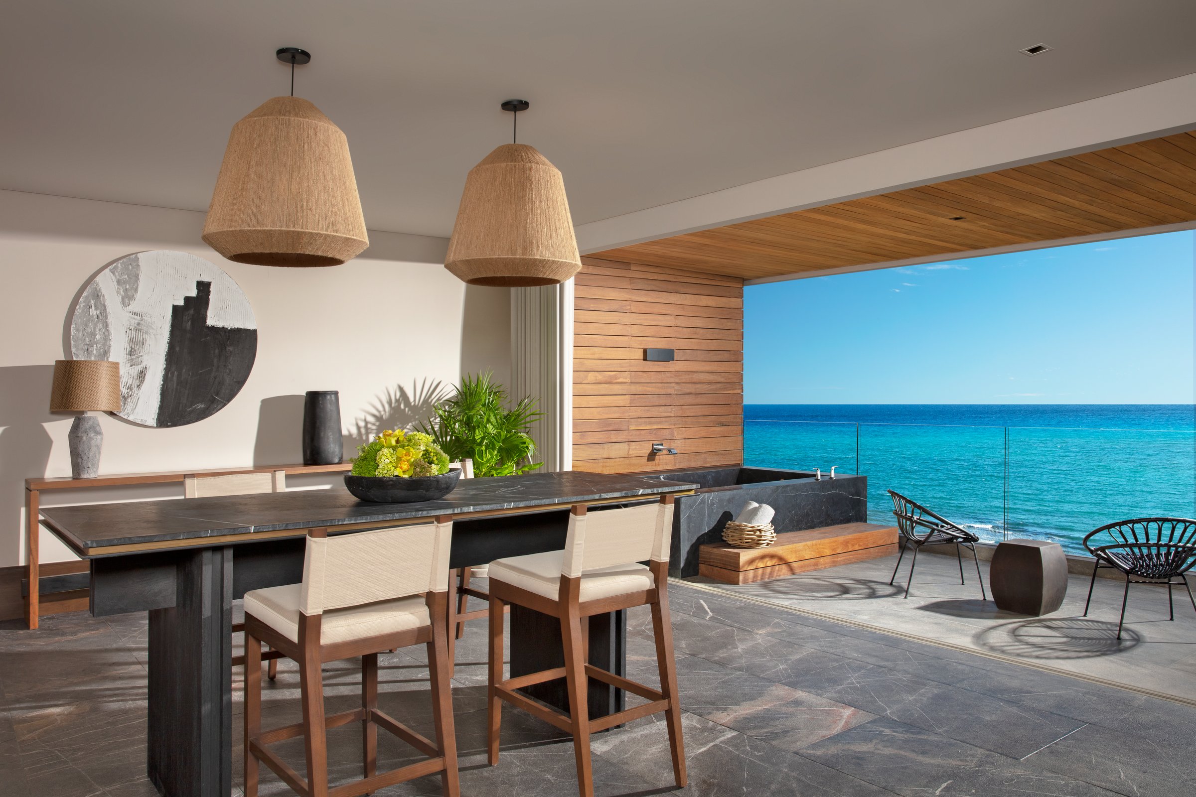 Suite dining area with a long wooden table under two wicker pendant lights, opening to a balcony and the turquoise ocean.