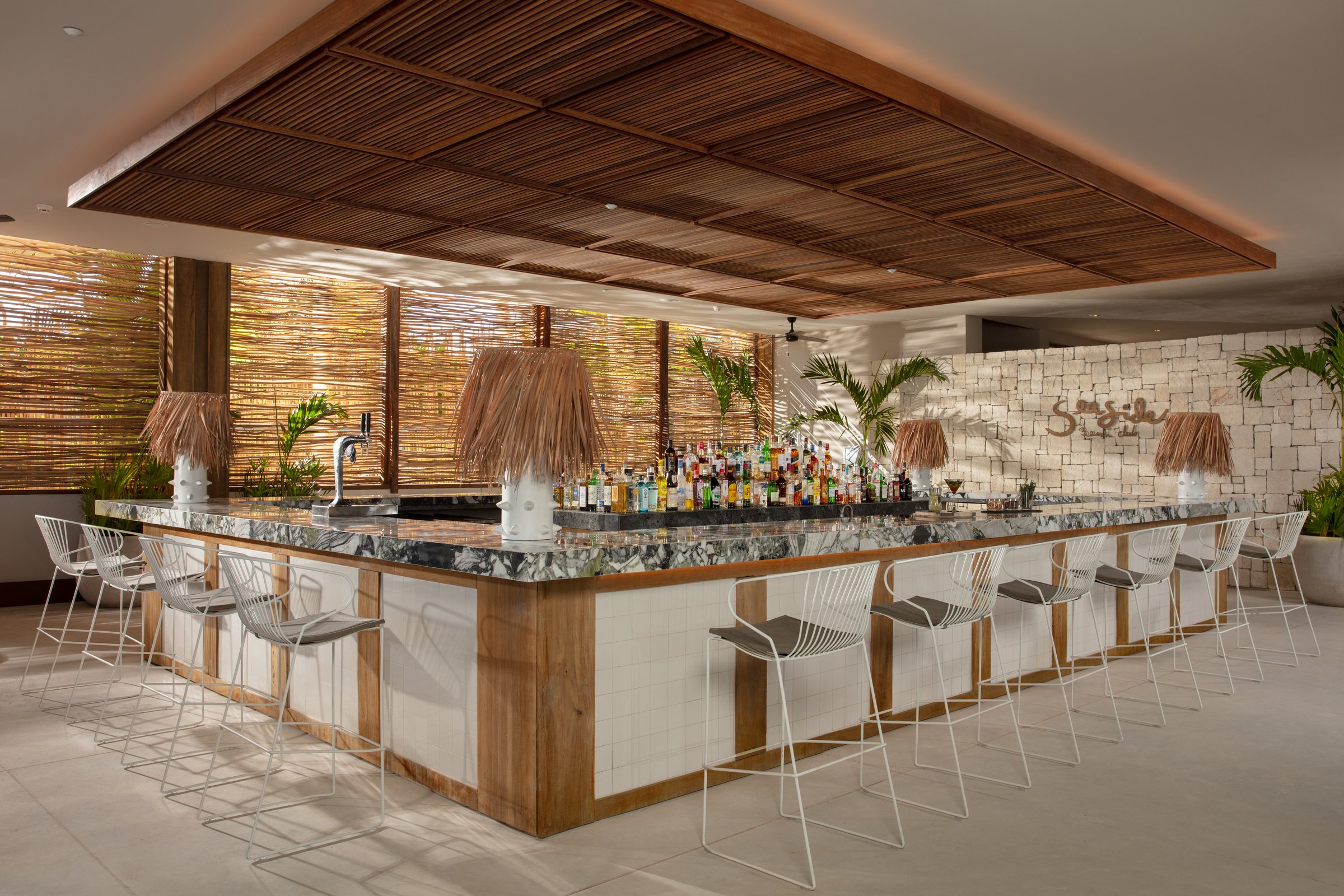 Seaside Beach Club open-air bar with a white tiled base, marble top, light wire stools and a woven branch ceiling.