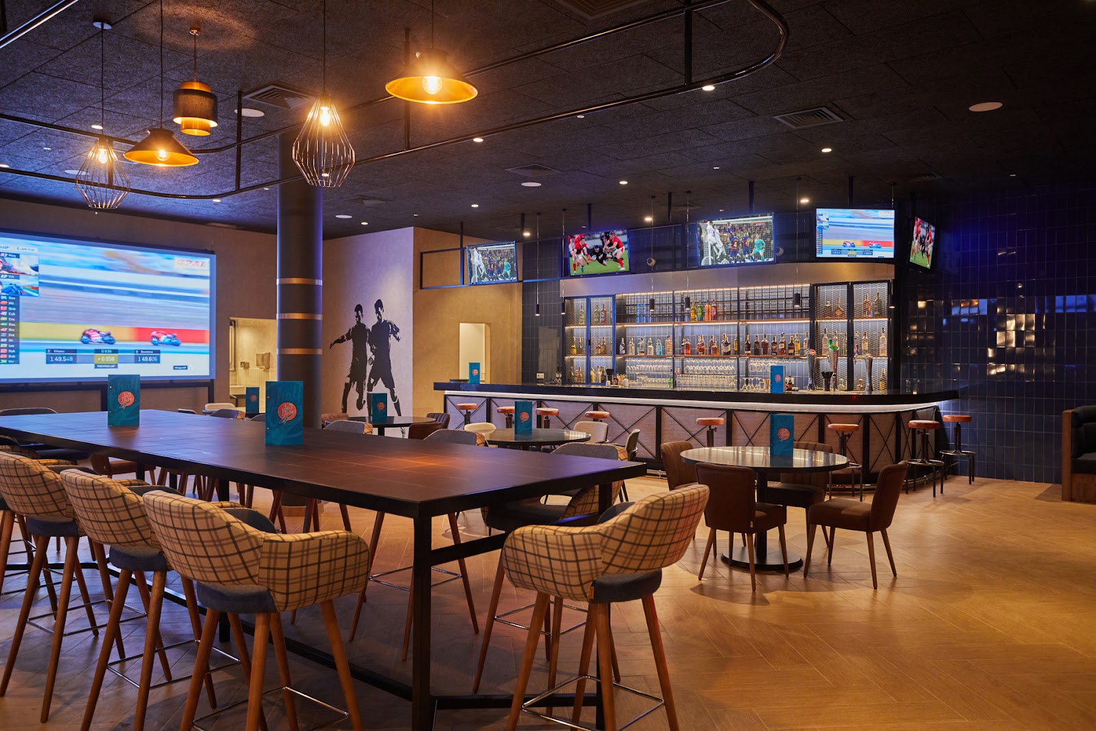 Sports bar with high-top tables, plaid upholstered chairs, illuminated liquor shelves, multiple TV screens, and pendant lighting overhead at Secrets Lanzarote Resort & Spa.