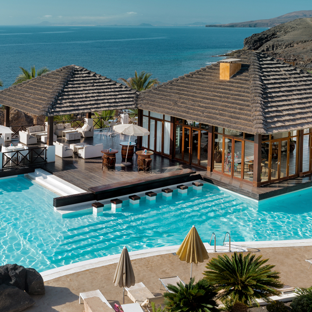 Turquoise pool with sunbeds and umbrellas overlooking the ocean, flanked by thatched-roof bungalows and wooden decking at Secrets Lanzarote Resort & Spa.