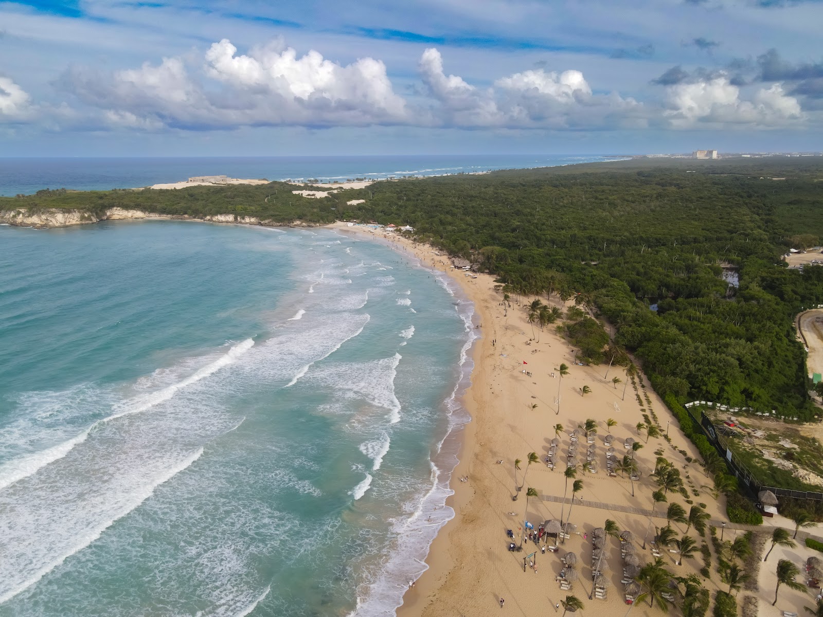 Aerial view of Secrets Macao Beach Punta Cana's sandy shore with waves rolling in, palm trees, sunbeds, and lush vegetation under a cloudy sky.