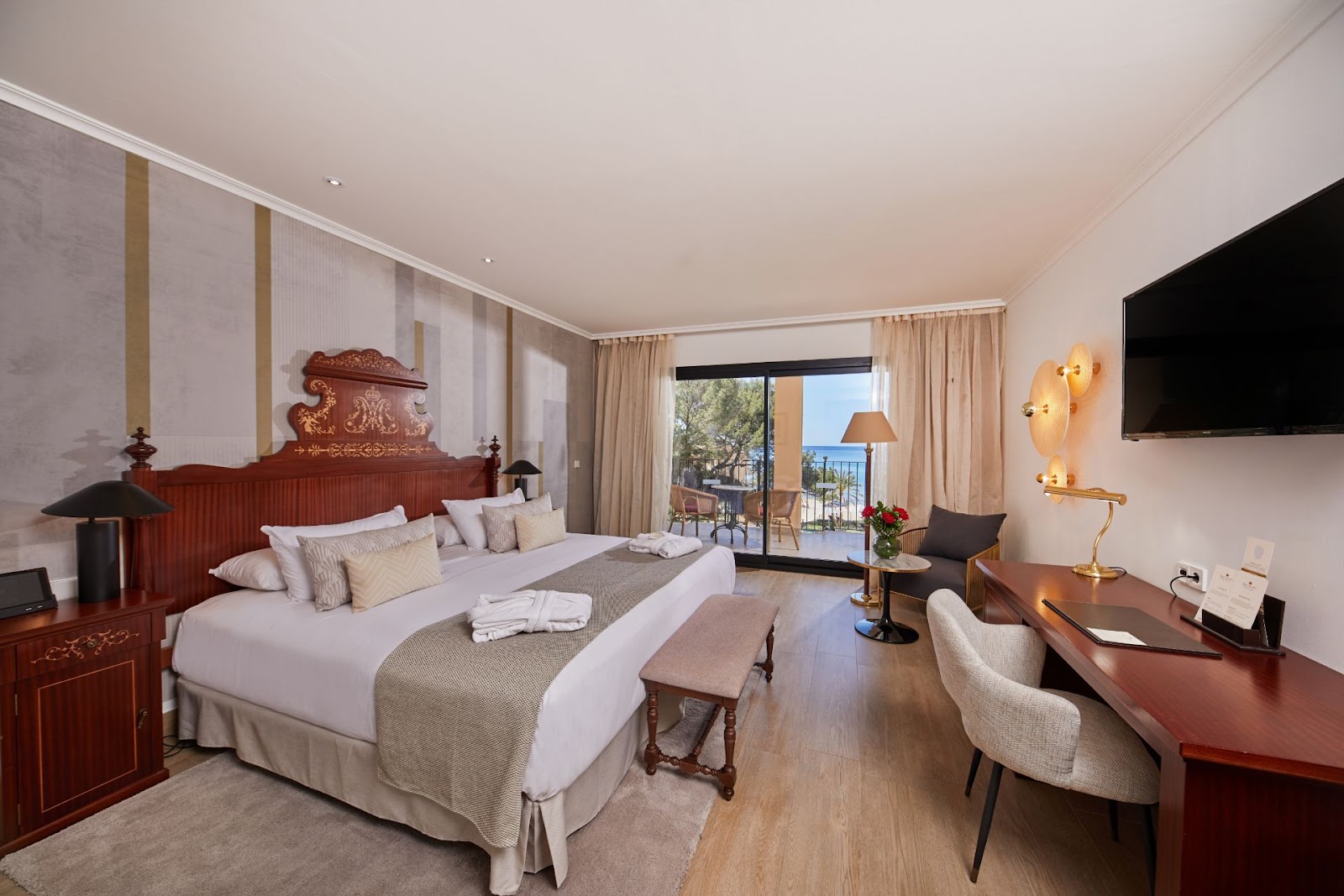 Upscale hotel bedroom with wooden furnishings, white bedding, and a balcony overlooking the Mediterranean coastline.