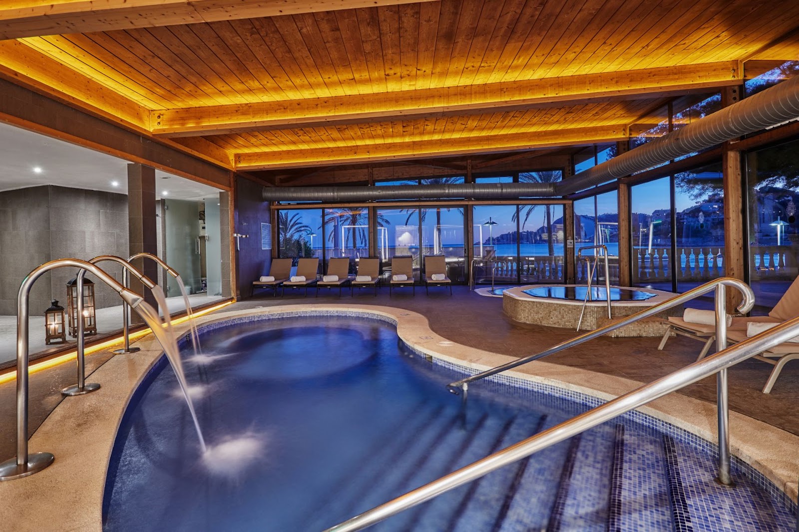 Indoor spa area with multiple blue thermal pools, wooden ceiling with warm lighting, and ocean views through floor-to-ceiling windows at dusk.