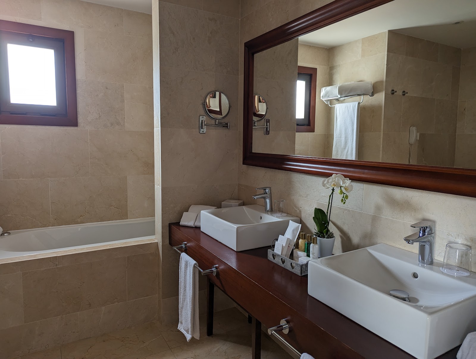 Resort bathroom with dual white vessel sinks on dark wood vanity, large mirror, soiled towels, orchid plant, and bathtub visible on left.
