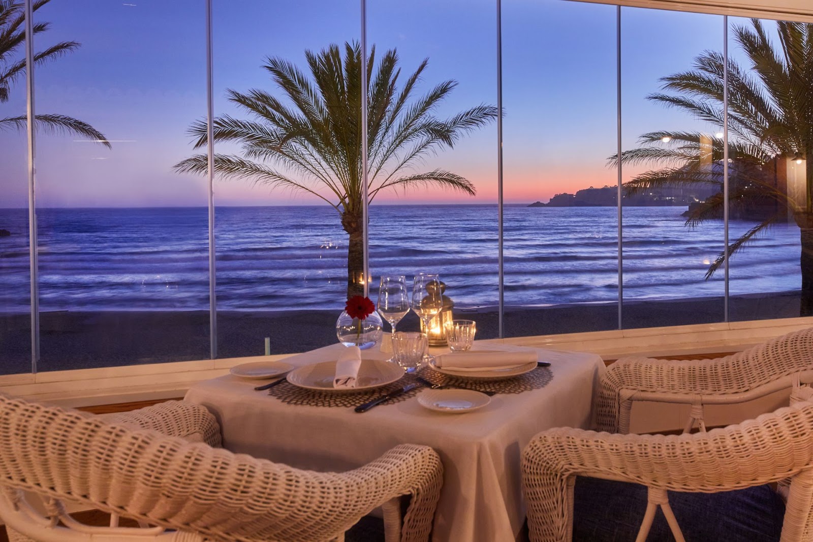 Beachfront dining room with wicker chairs, set table, and ocean view at sunset with palm tree and candlelit glassware.