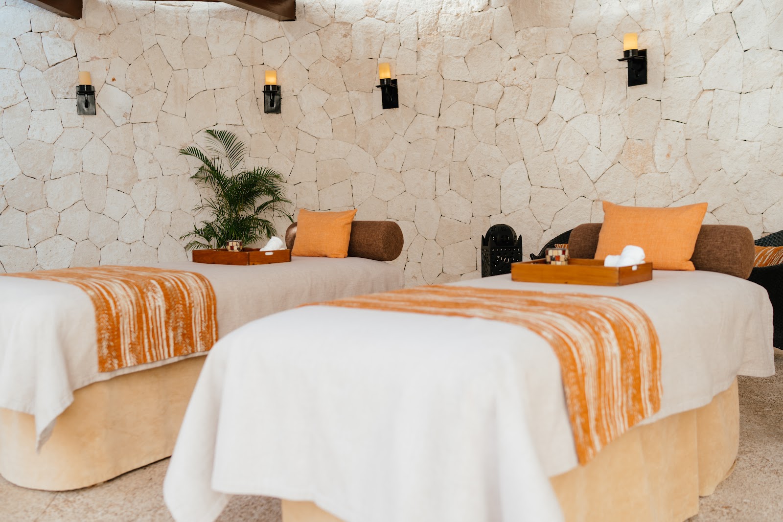 Spa treatment room with two massage tables draped in white linens and orange striped blankets, cream stone wall, wall-mounted torch sconces, and brown accent pillows at Secrets Maroma Beach Riviera Cancun.