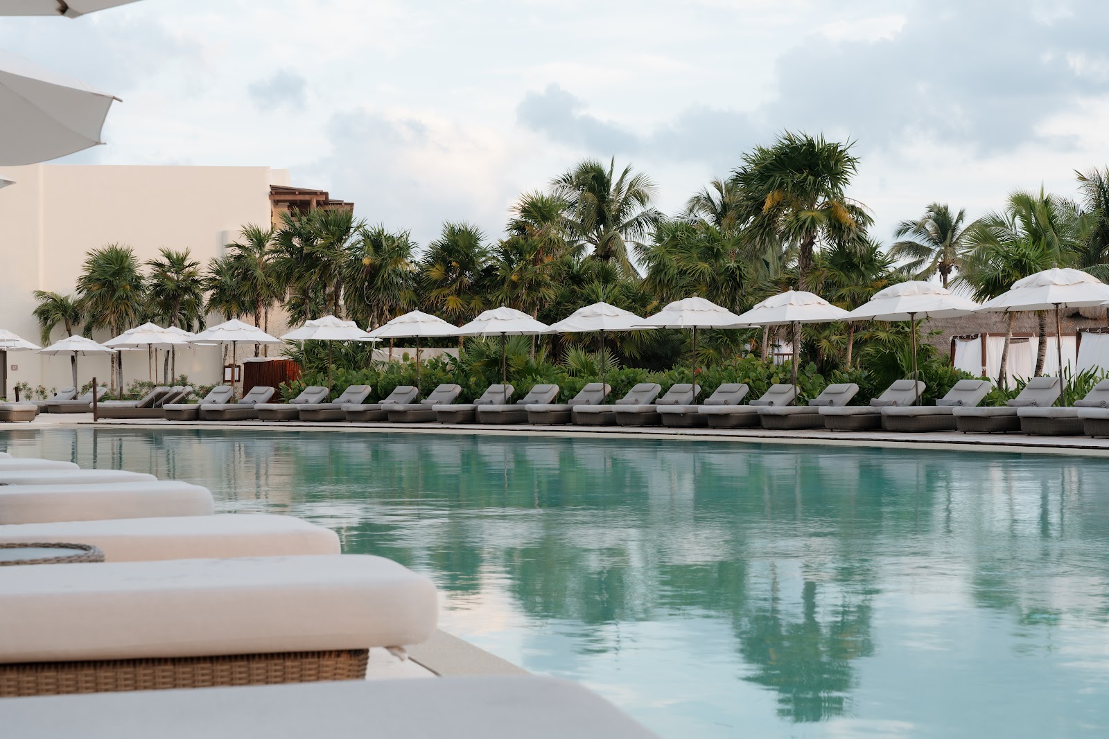 Turquoise pool lined with white umbrellas and lounge chairs, palm trees and resort buildings in background under cloudy sky.