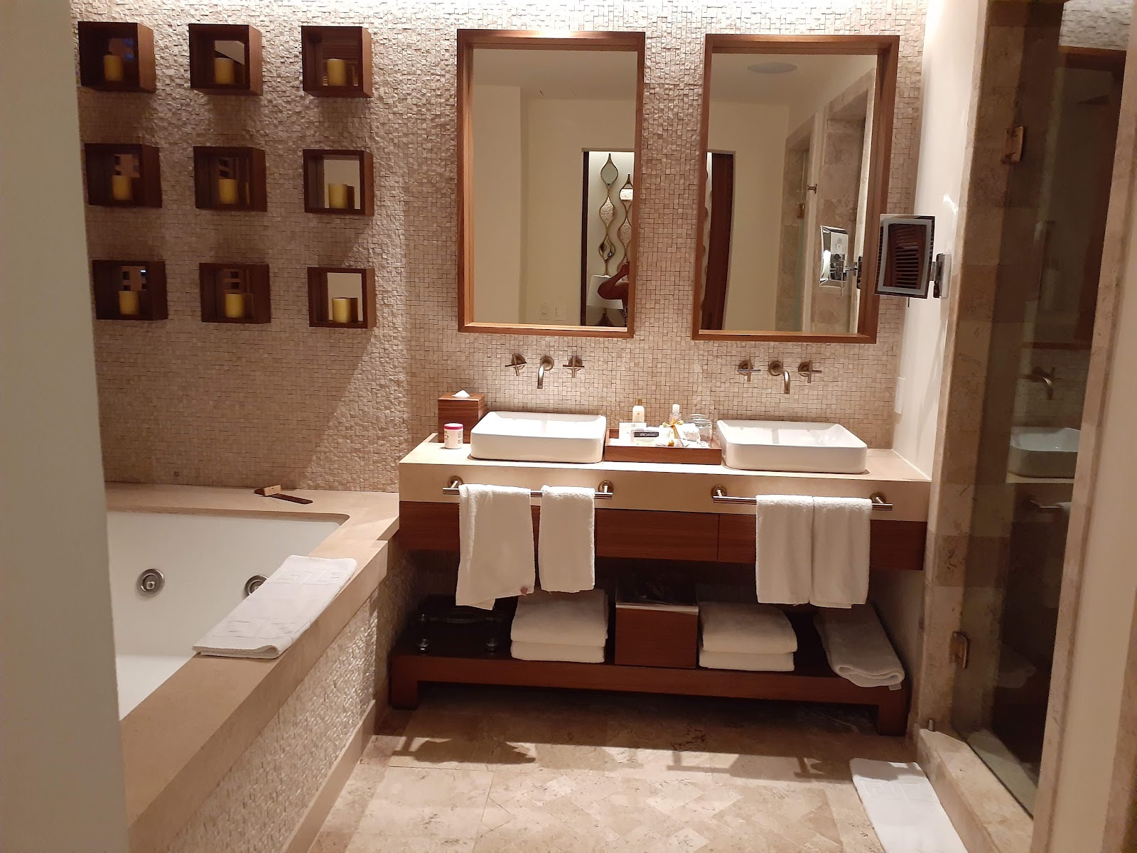 Spa-style bathroom with dual vessel sinks, large mirrors, soaking tub, cream tile walls, and wooden niche shelving at Secrets Maroma Beach Riviera Cancun.