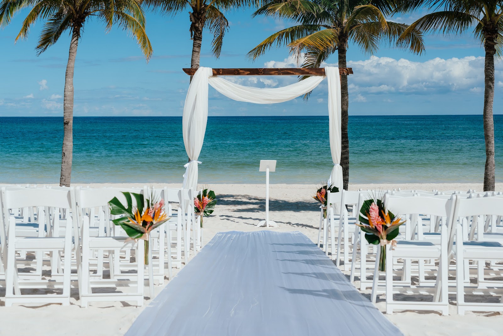 Beach wedding ceremony setup with white chairs, tropical flowers, and draped archway facing turquoise ocean and palm trees.