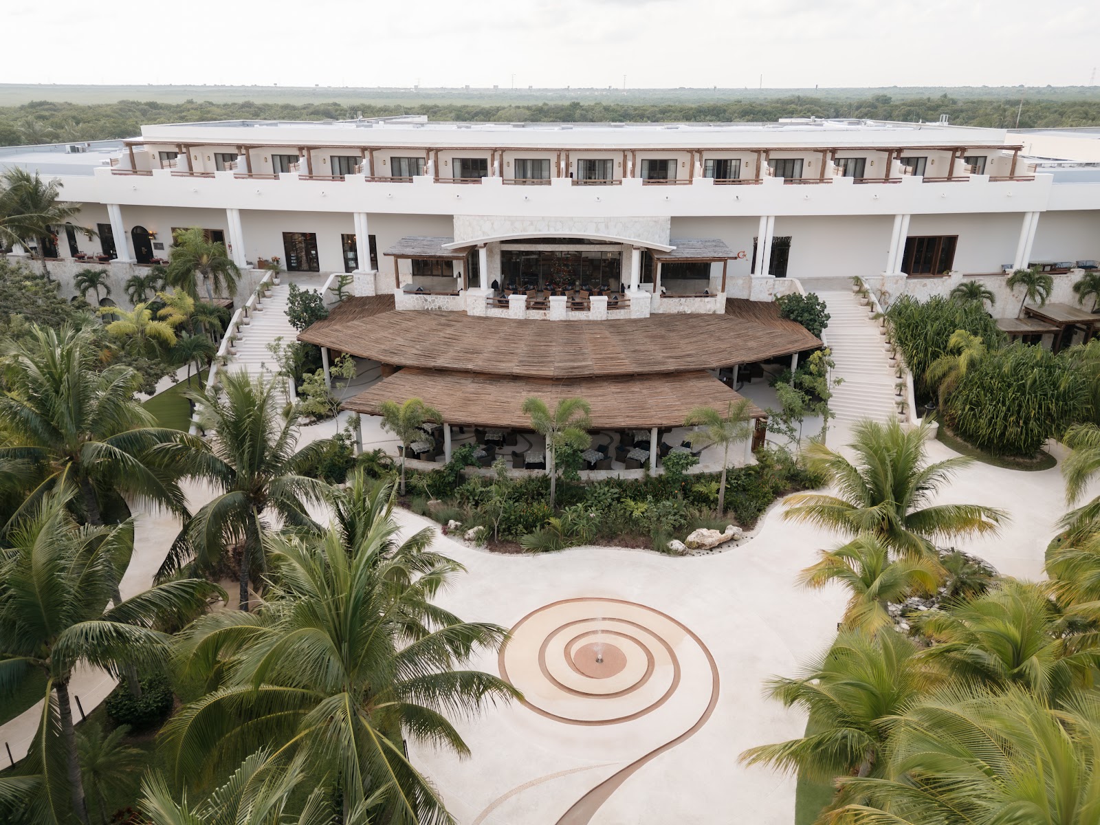 Aerial view of Secrets Maroma Beach Riviera Cancun's white courtyard with tiered wooden palapas, spiral paving design, and palm trees, with the waterfront visible beyond.