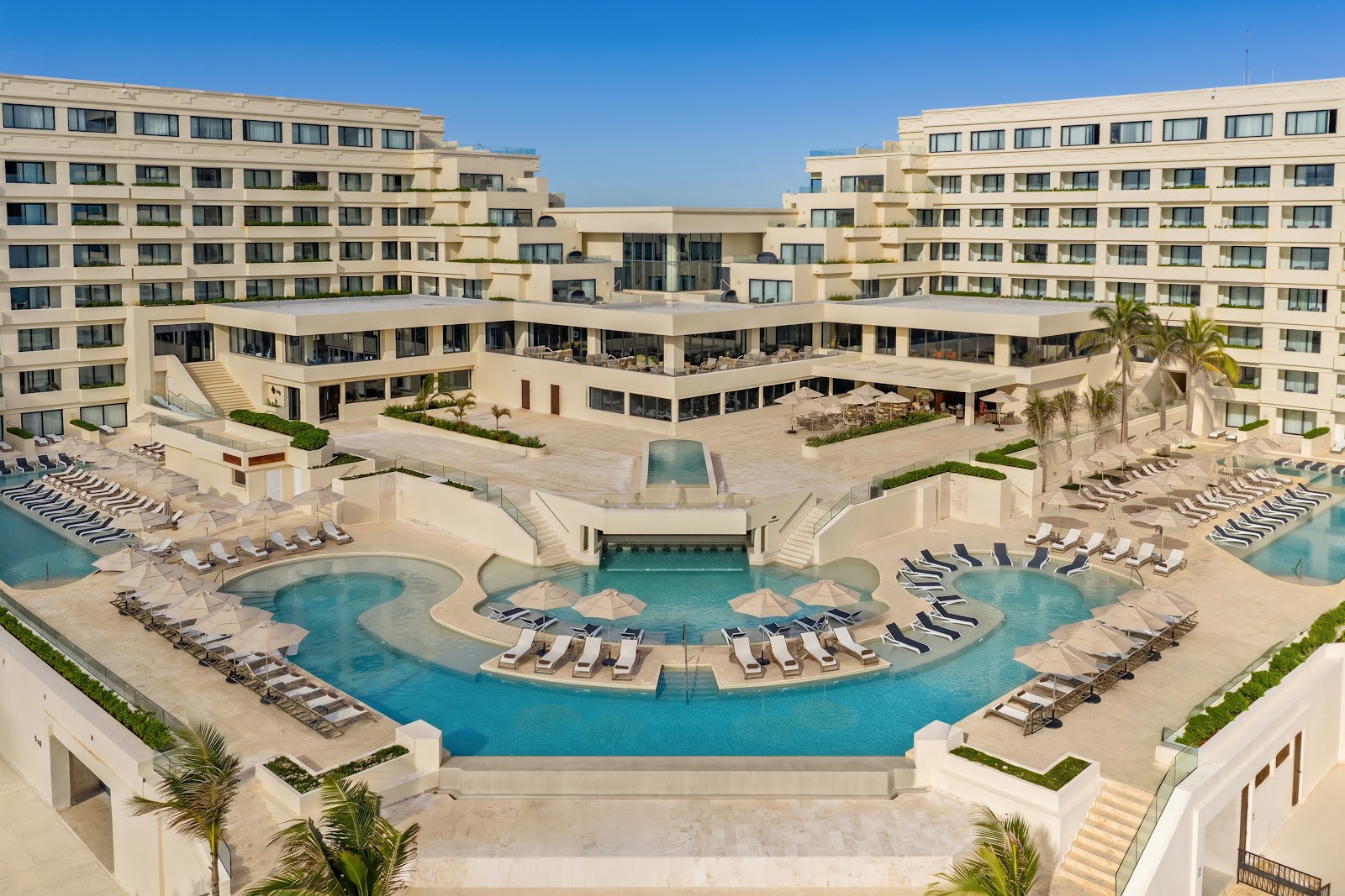 Beige resort buildings surround a large curved pool with lounge chairs and umbrellas, palm trees, and ocean beach area under clear blue sky.