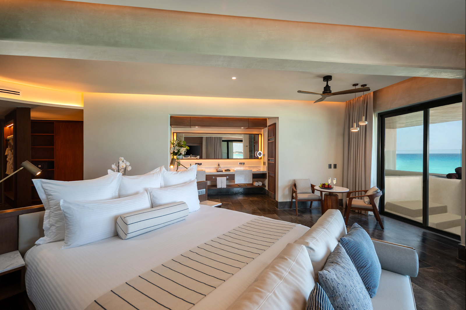 Spacious resort bedroom with king bed, ensuite bathroom with lit mirror, seating area, and ocean view through sliding glass doors.