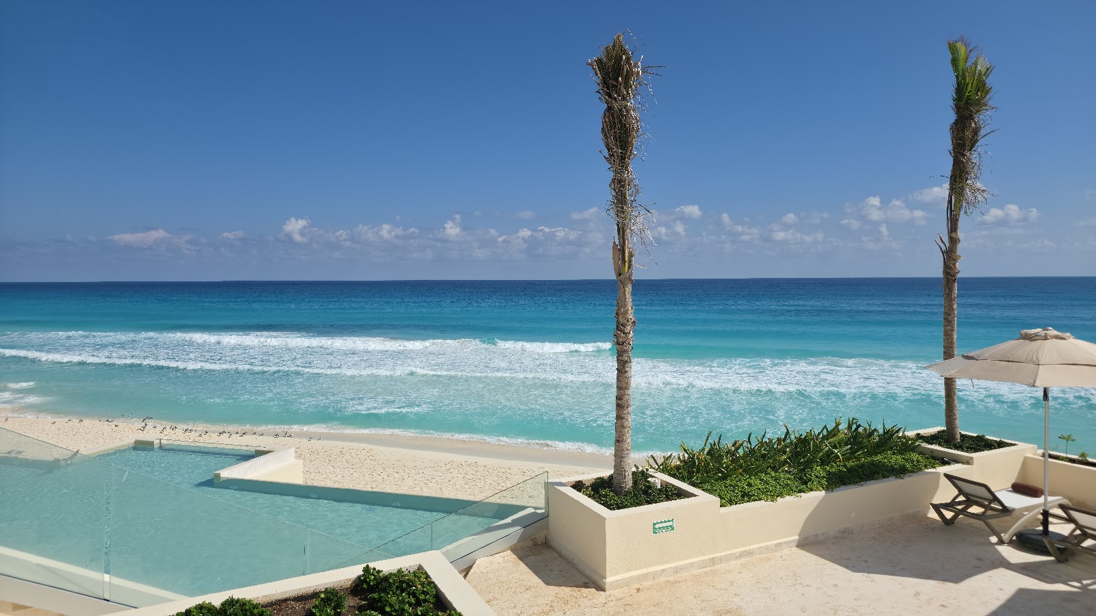 Beachfront pool terrace with turquoise water, white sand, palm trees, and ocean horizon under clear sky at Secrets Mirabel Cancun.