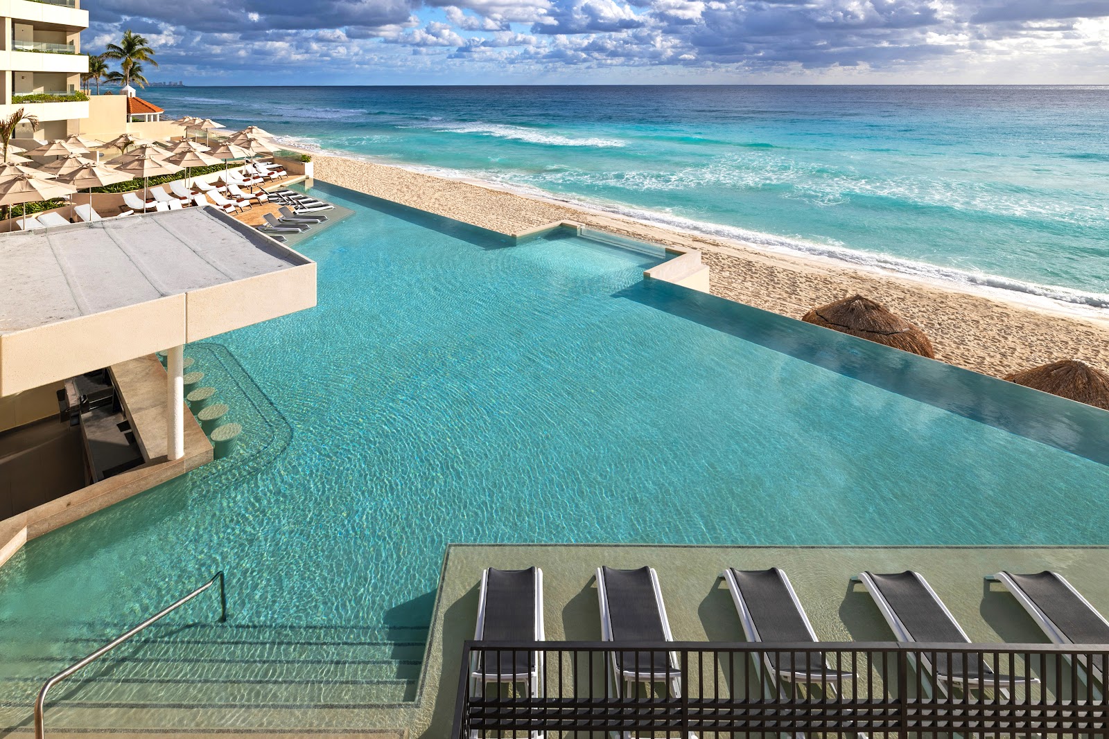 Resort infinity pool overlooking turquoise Caribbean waters and sandy beach with loungers and umbrellas.