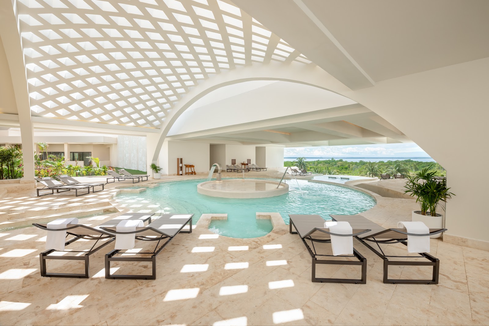 Indoor pool with loungers and towels beneath latticed ceiling, central hot tub, and ocean view at Secrets Mirabel Cancun.
