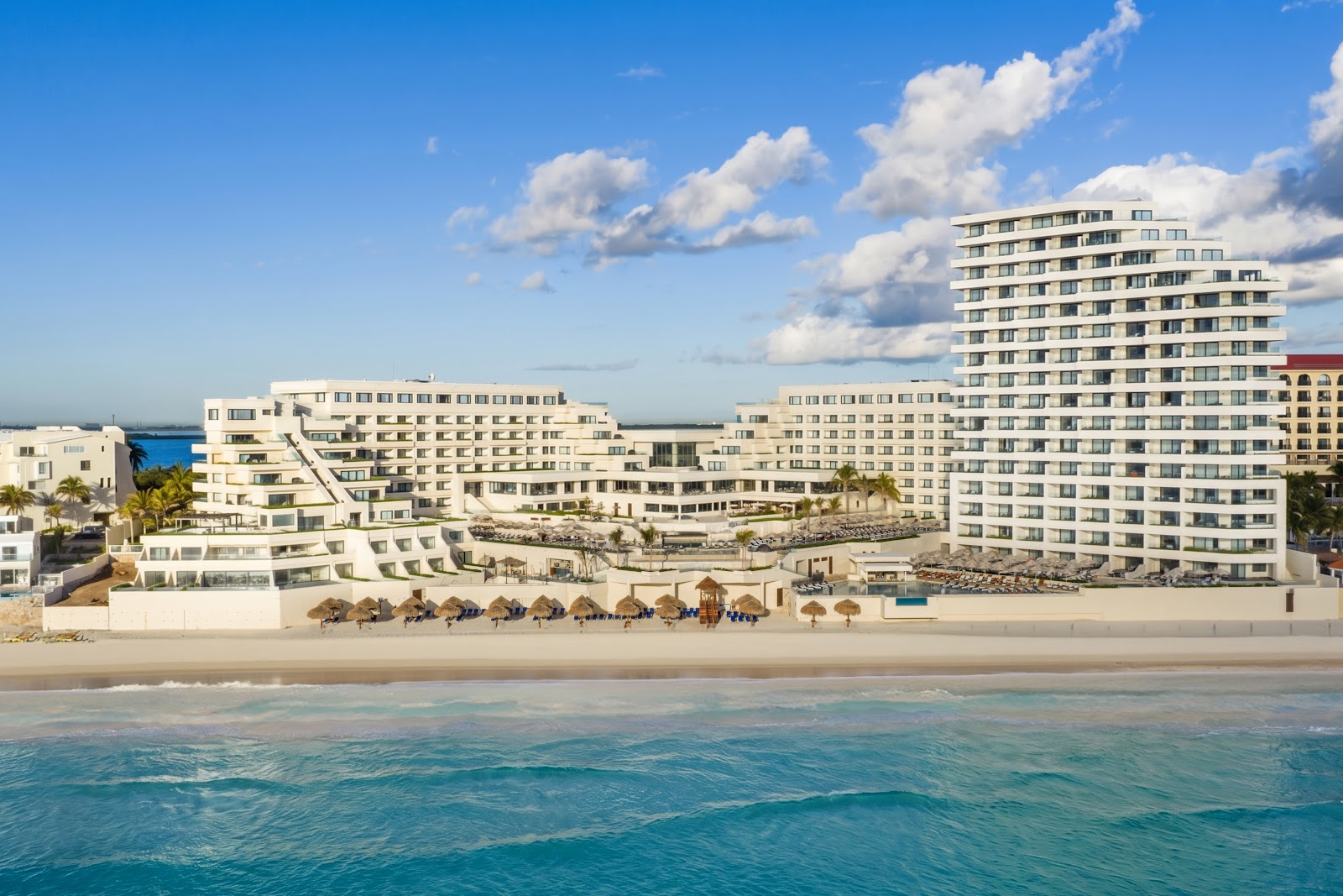 White multi-story resort buildings line a sandy beach with turquoise water and thatched umbrellas along the shore at Secrets Mirabel Cancun.