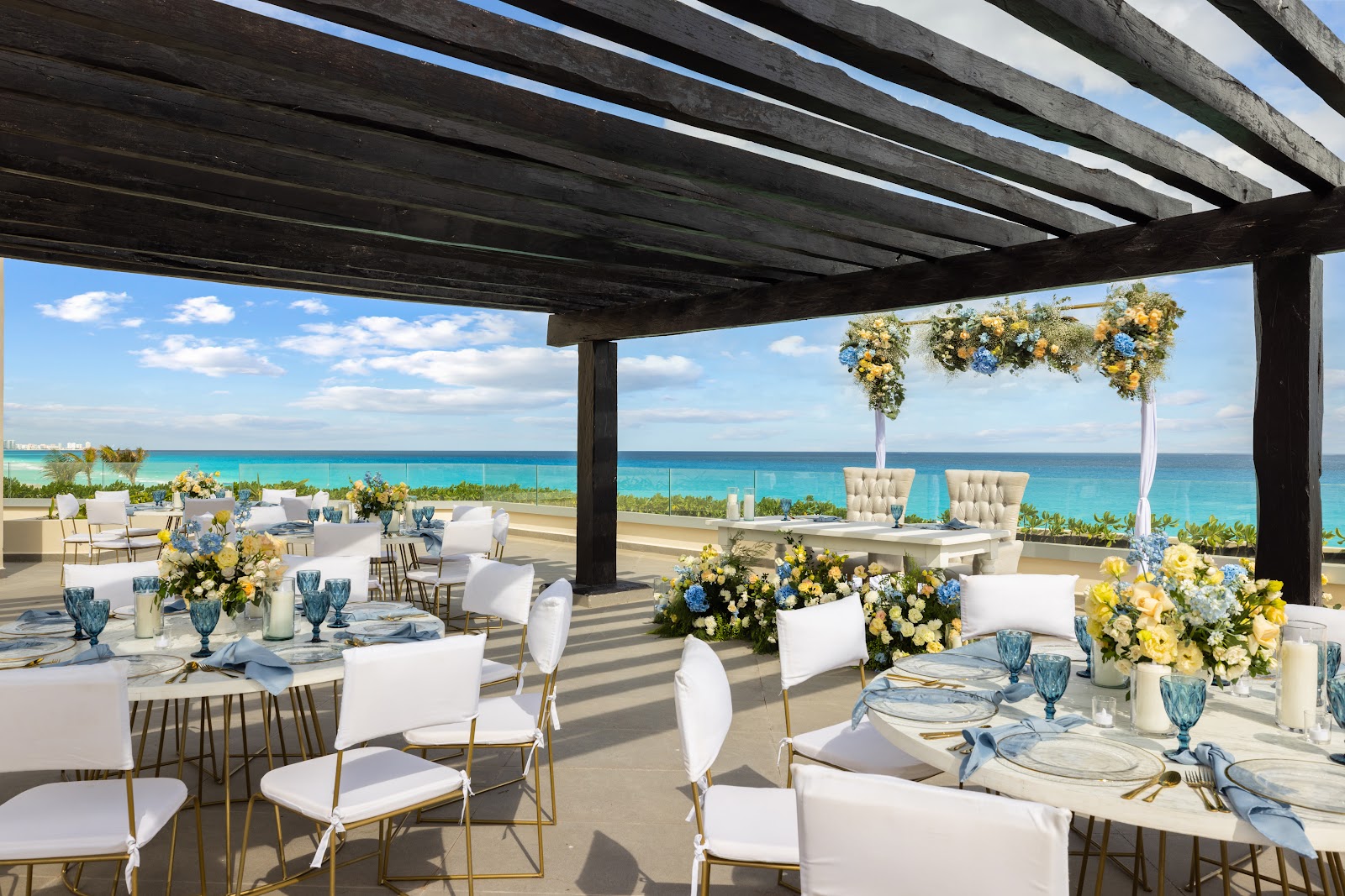 Beachfront terrace at Secrets Mirabel Cancun with turquoise ocean view, white dining tables, yellow and blue floral arrangements, and dark wooden pergola overhead.