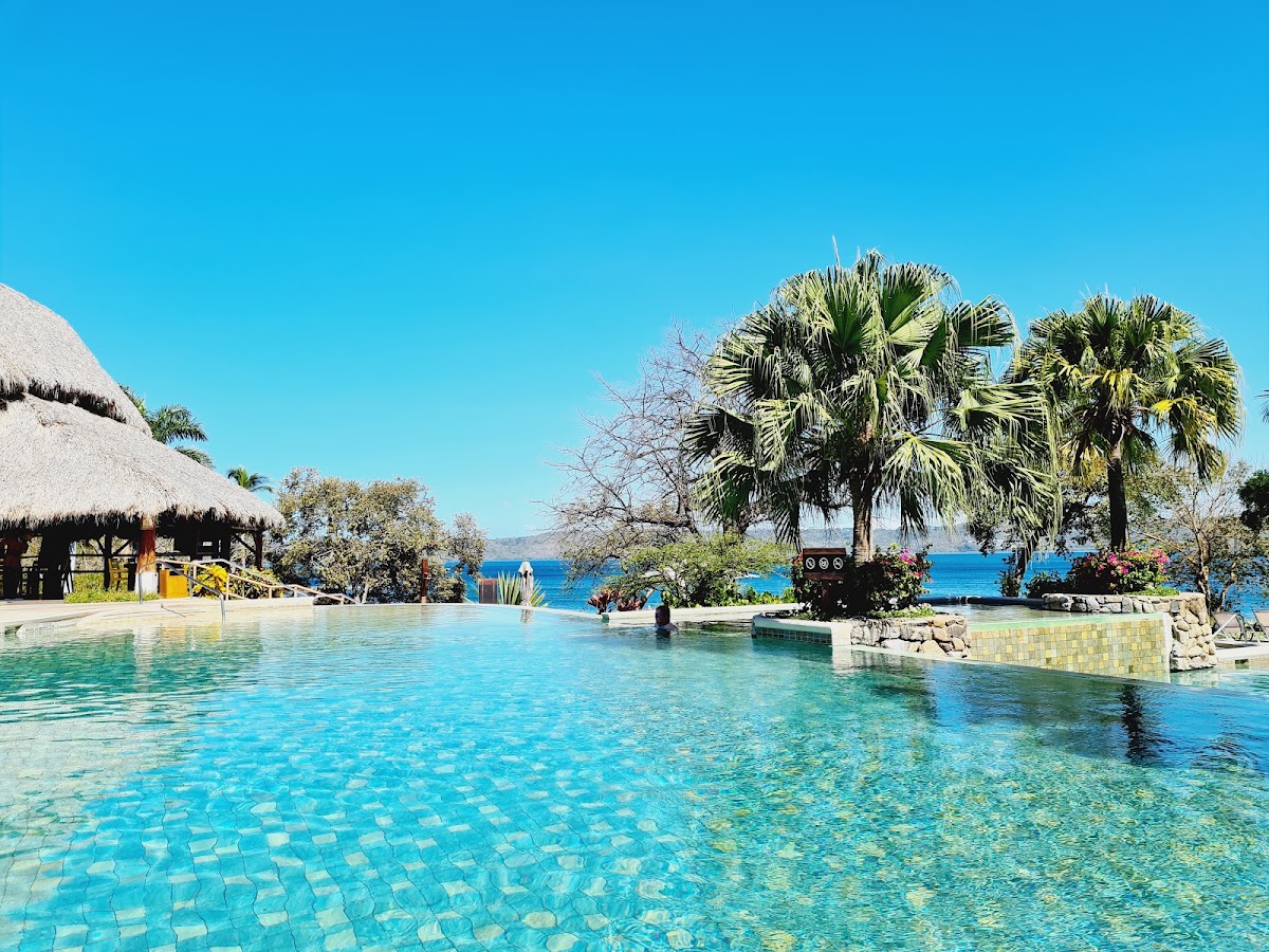 Resort pool with thatched-roof bar, palm trees, and clear ocean view under blue sky at Secrets Papagayo Costa Rica.