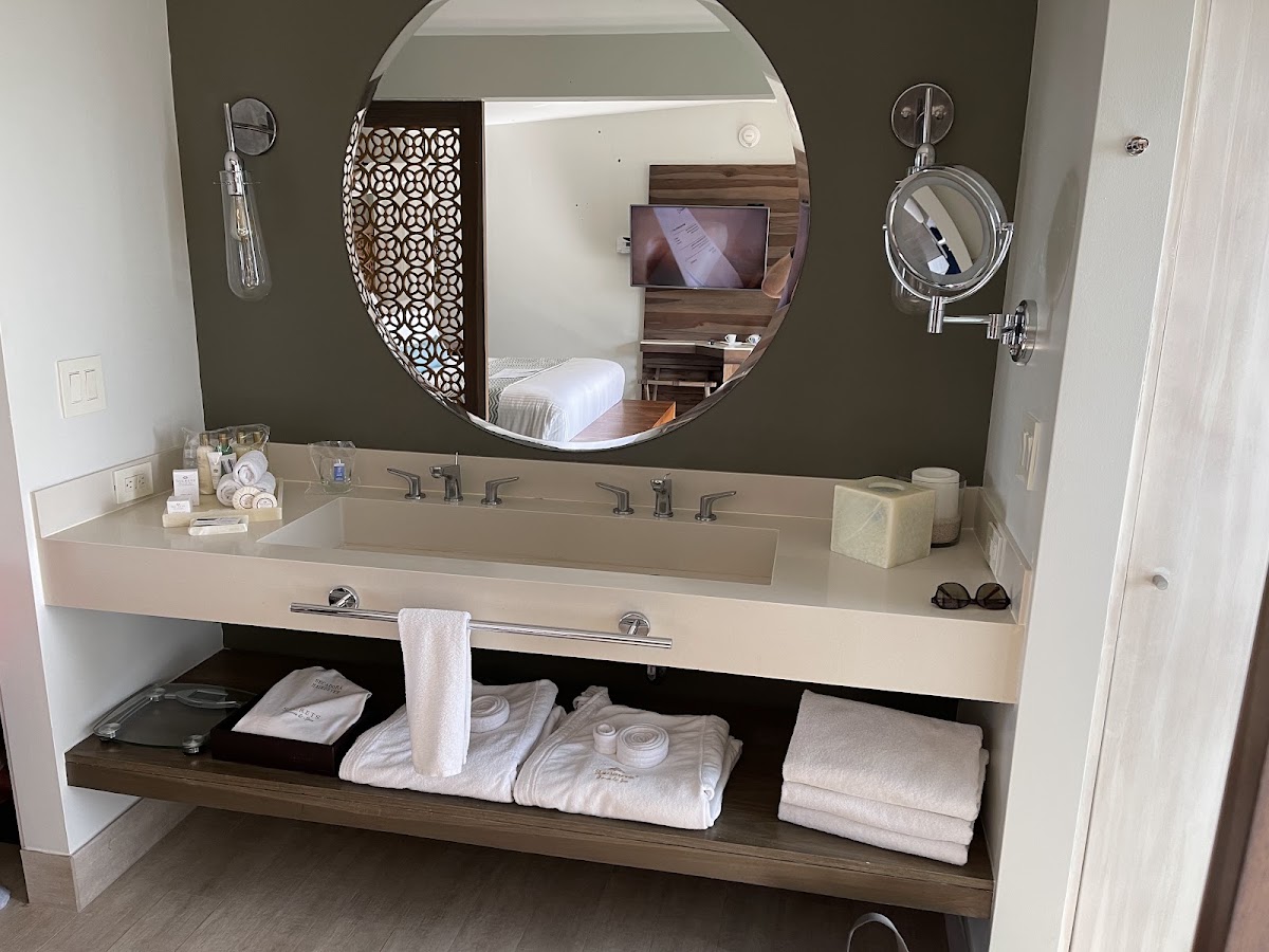 Modern bathroom vanity with long countertop sink, round mirror, and open shelving holding white towels and toiletries, bedroom visible in mirror reflection.