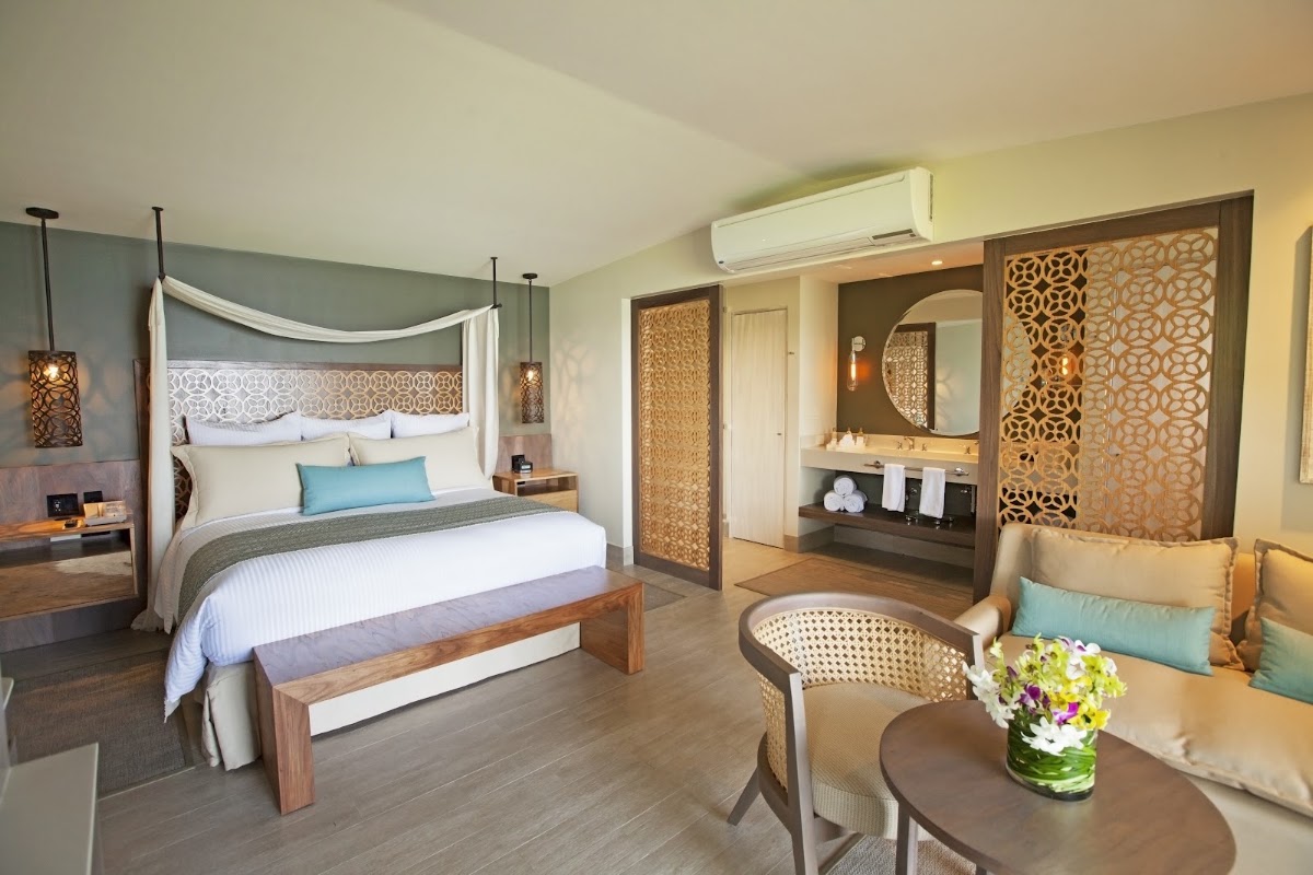 Spacious resort suite with four-poster canopy bed, ensuite bathroom with round mirror, sitting area with flowers, and geometric wood screens throughout.