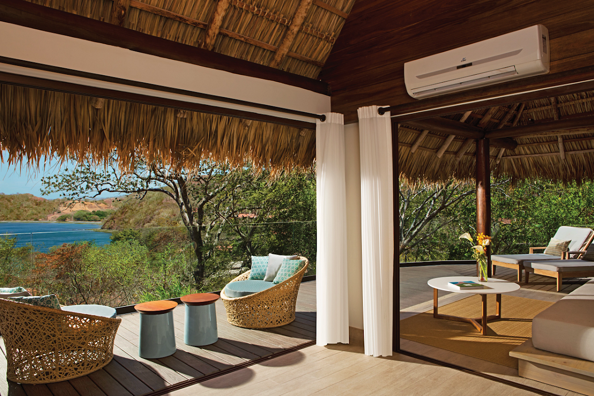 Thatched-roof terrace at Secrets Papagayo Costa Rica with woven chairs, ocean view, and lush hillside vegetation beyond.