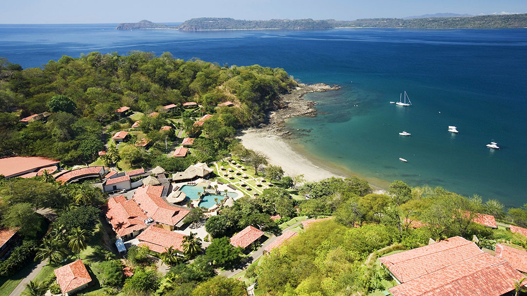 Aerial view of Secrets Papagayo Costa Rica nestled on a forested peninsula with a pool, beach, and sailboats anchored in turquoise water.