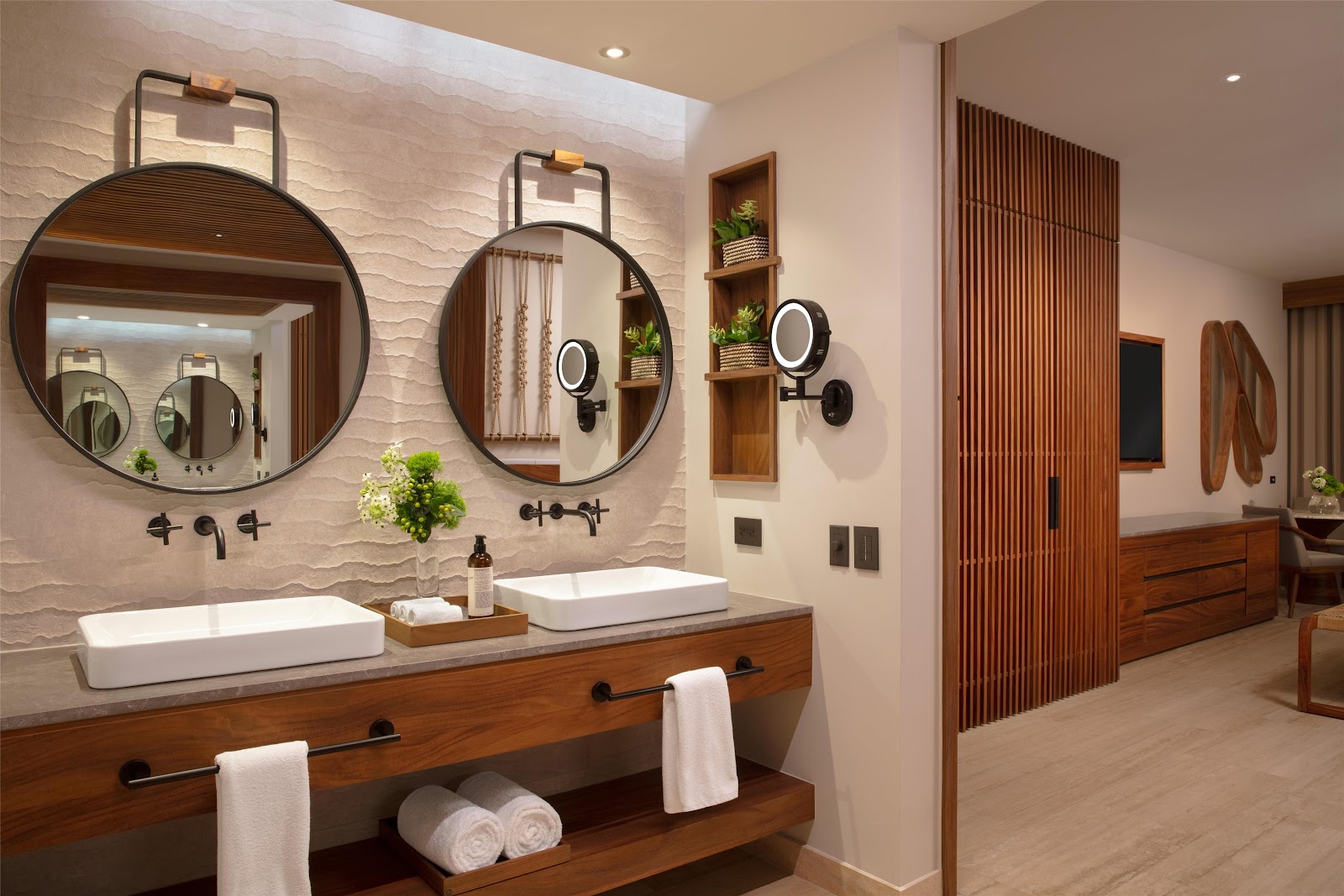 Bathroom with dual wooden vanity, two round mirrors with black frames, white rectangular sinks, textured wall, and potted plants on shelving.