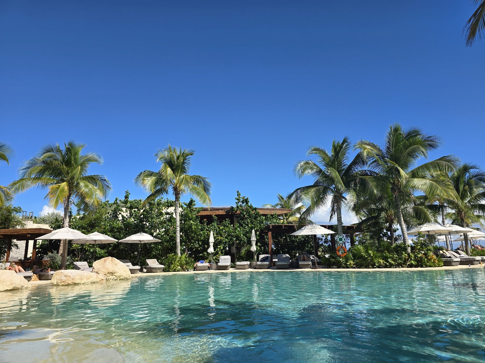 Turquoise pool at Secrets Playa Blanca Costa Mujeres with lounge chairs, white umbrellas, palm trees, and multi-story resort buildings under blue sky.