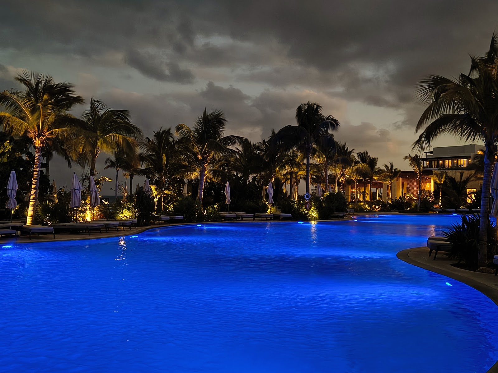 Curved resort pool with turquoise water overlooks sandy beach, thatched-roof palapas, and ocean under partly cloudy sky.