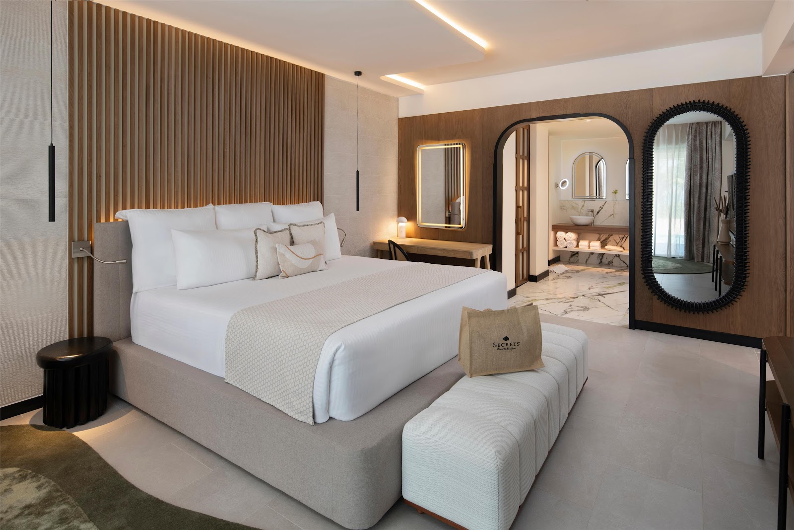 Modern resort bedroom with king bed, white linens, wood accent wall, ensuite bathroom visible through arched doorway, and decorative ottomans.